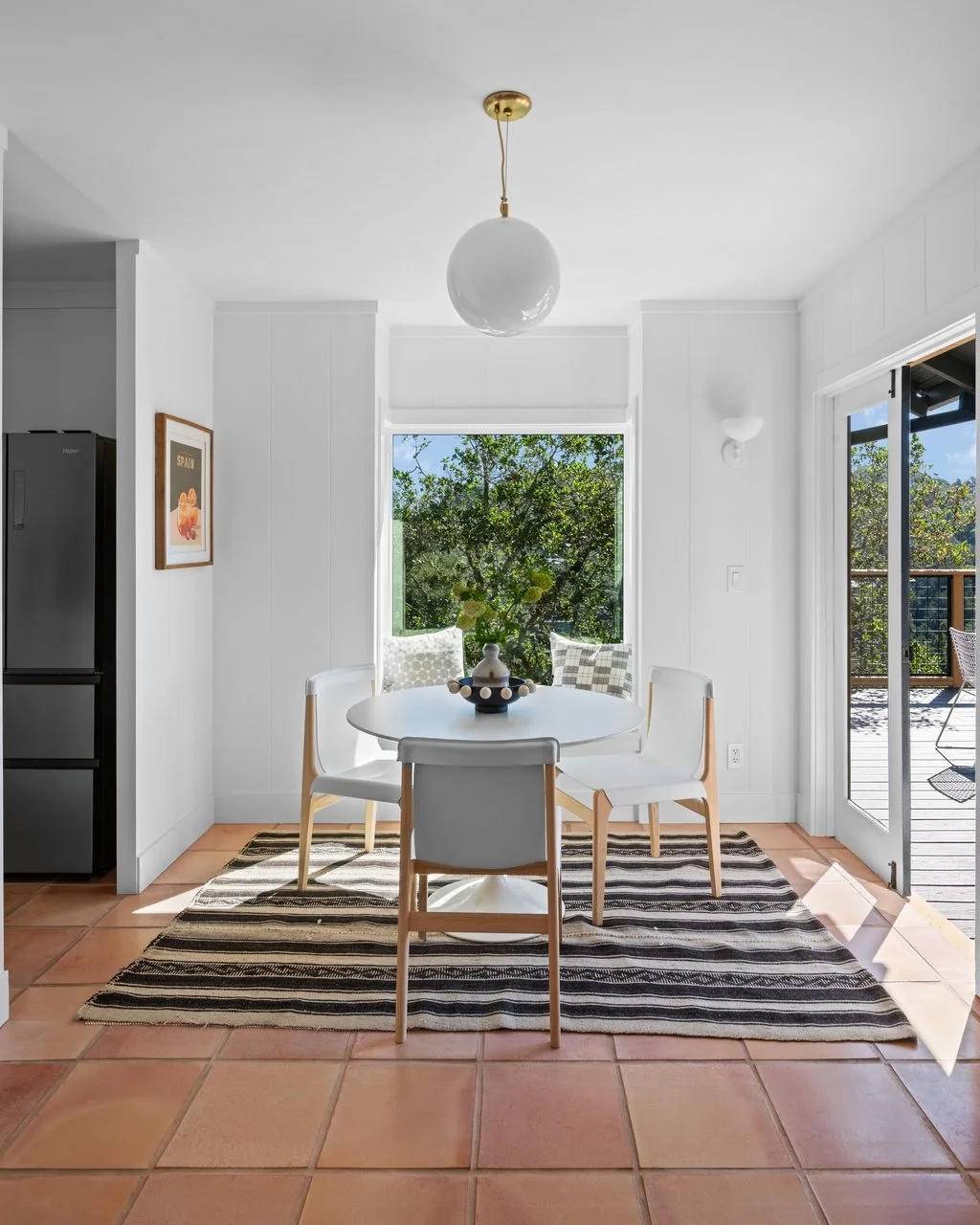 Bright dining area with a round white table, four white chairs, a black and white striped rug, a window with outside greenery, and a sliding glass door leading to an outdoor deck.