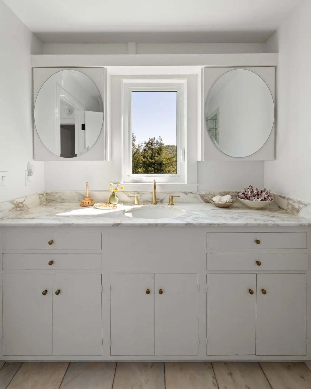 A bathroom vanity with a marble countertop, two oval mirrors, a window showing trees outside, and decorative items like a soap brush, a vase with flowers, and shells.