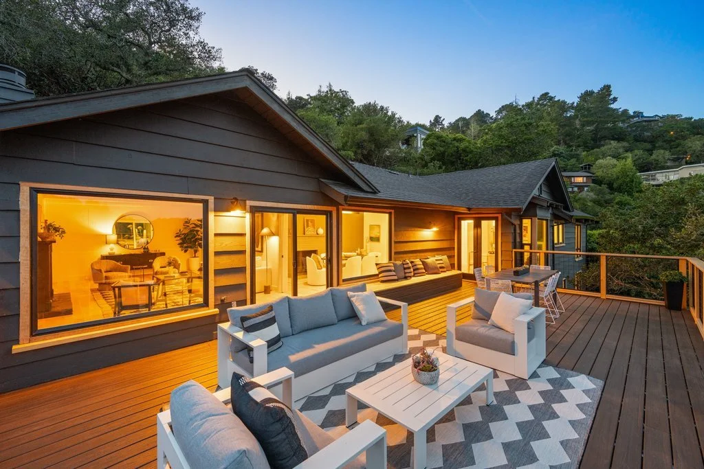 Outdoor wooden deck at dusk with modern seating, including a sofa and chairs with cushions, a coffee table with a small plant, and a patterned rug. The house has large windows and sliding doors, revealing a warmly lit interior with more seating and g
