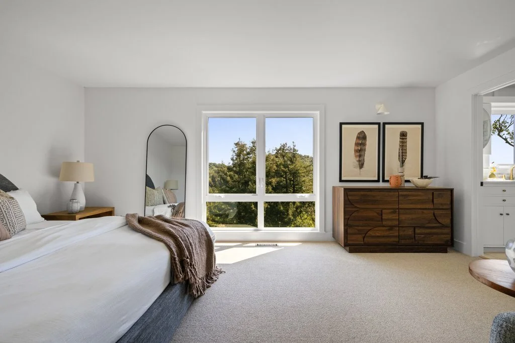 Bright bedroom with large window overlooking trees, white walls, bed with white bedding and brown throw blanket, round mirror, wooden nightstand with lamp, dark wood dresser with framed artwork of feathers above.
