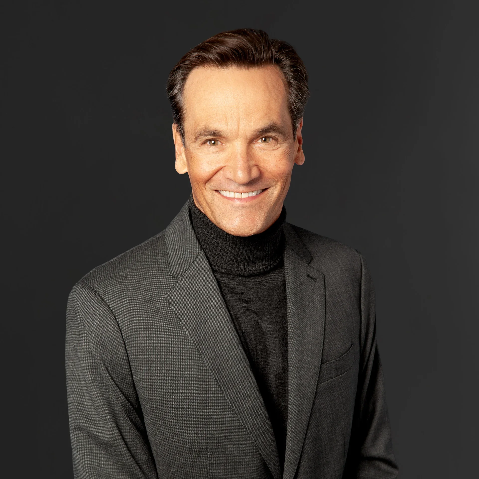 A smiling man in a gray suit with a black turtleneck against a dark background.