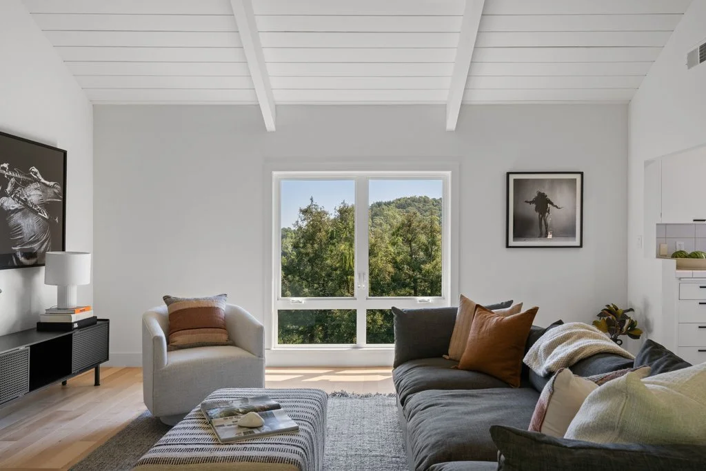 Bright living room with white walls, large window with outdoor greenery, gray sofa with pillows, white armchair, artwork on walls, and a dark entertainment unit.