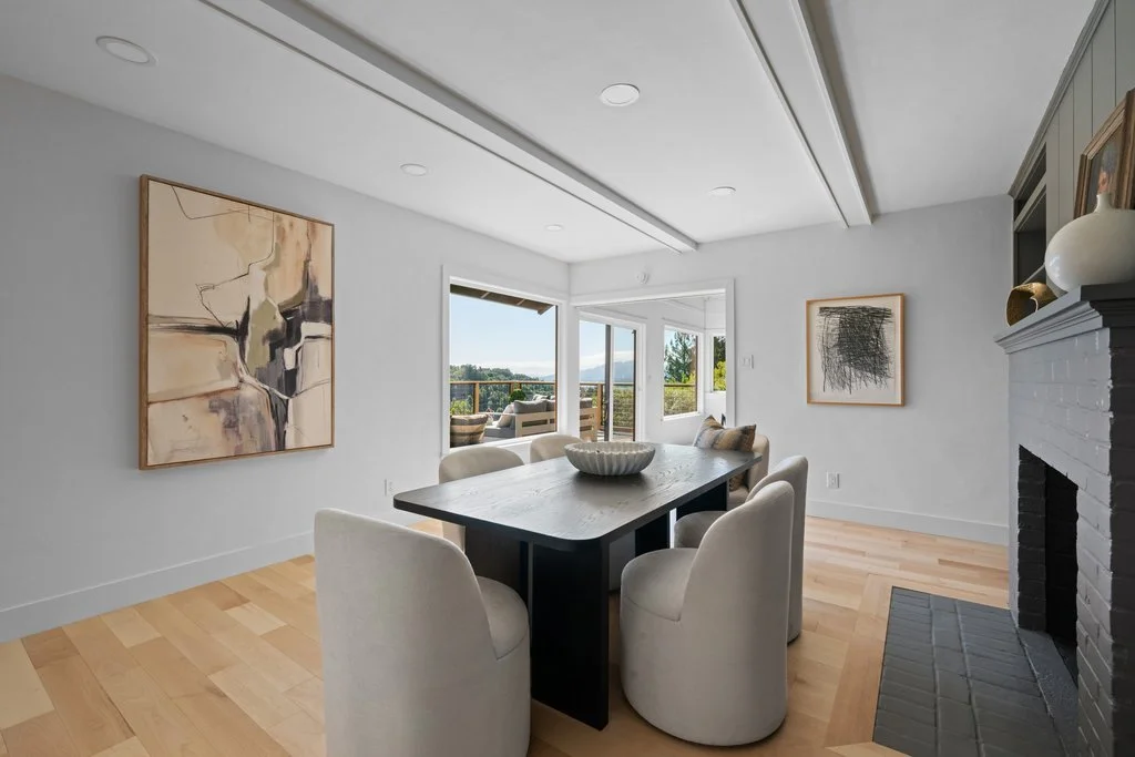 A dining room with a black rectangular table surrounded by six white upholstered chairs. Artwork hangs on the white walls, and a window opens to a balcony with outdoor seating, overlooking a scenic view.