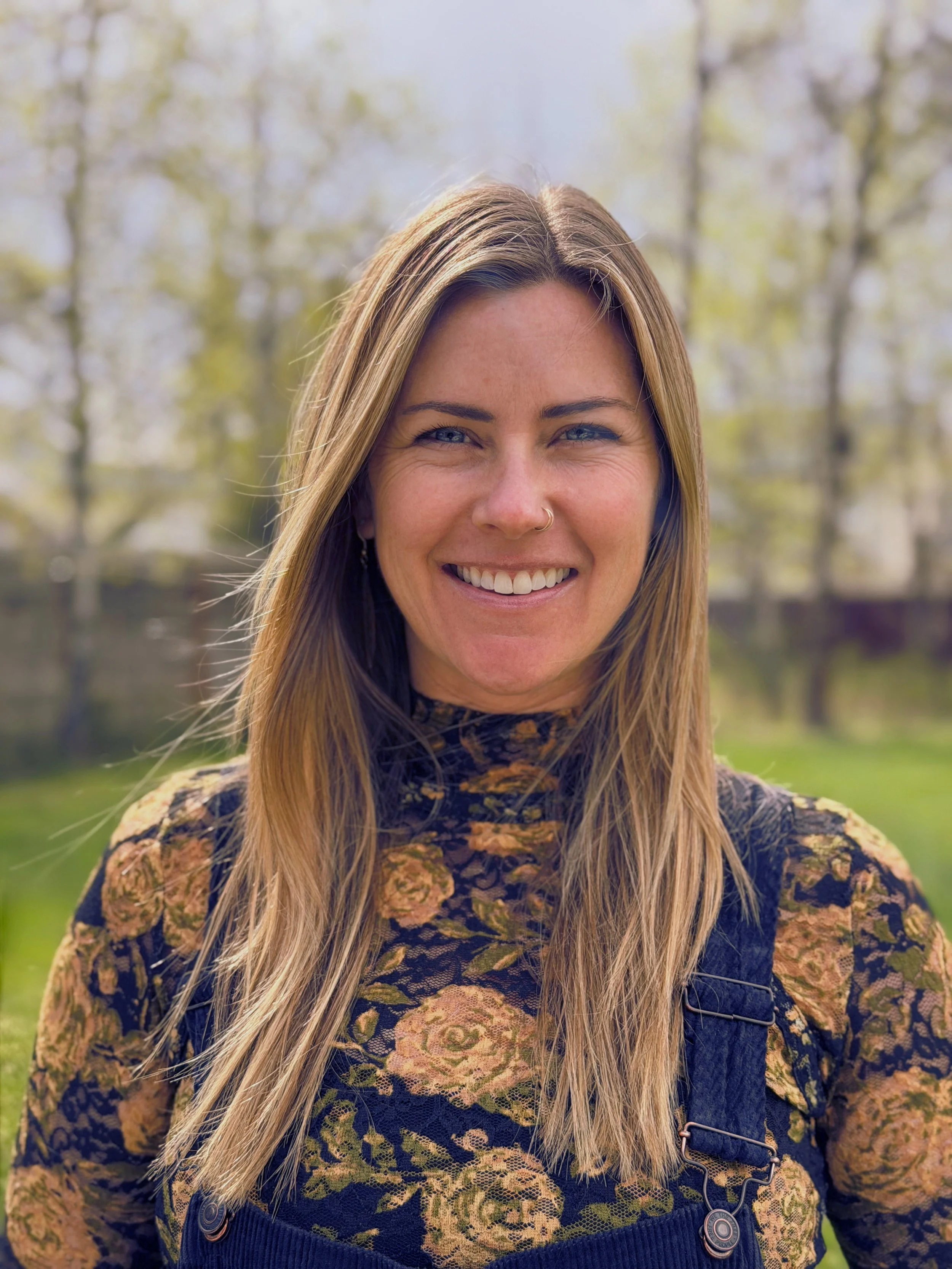 Close-up of a smiling woman with long blonde hair, wearing a floral-patterned top and a blue jumper, standing outdoors with blurred trees in the background.