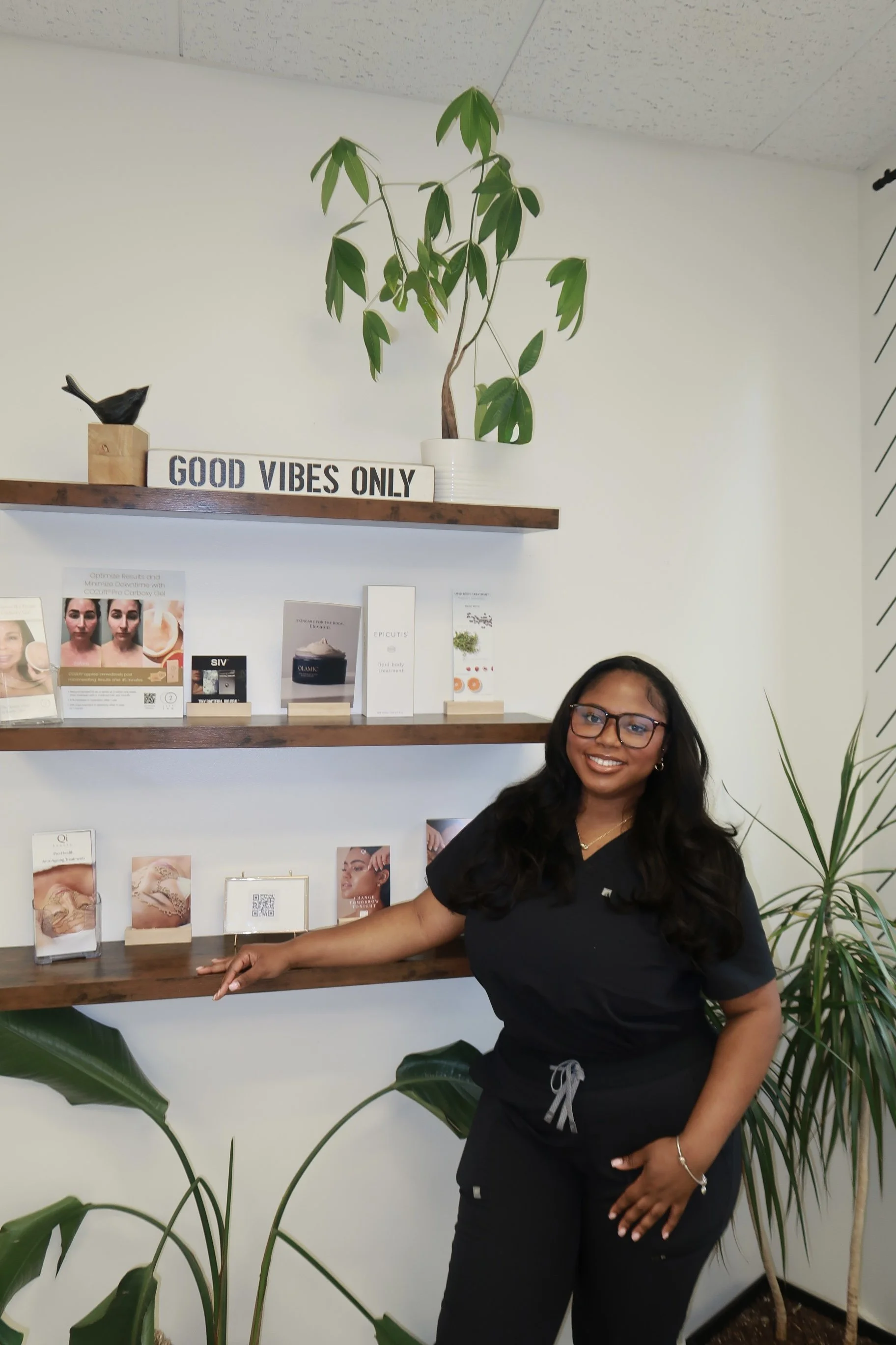 A woman with glasses and long dark hair, wearing black scrubs, standing in front of a white wall with wooden shelves displaying skincare and wellness products, plants, and a sign that says 'Good Vibes Only'.