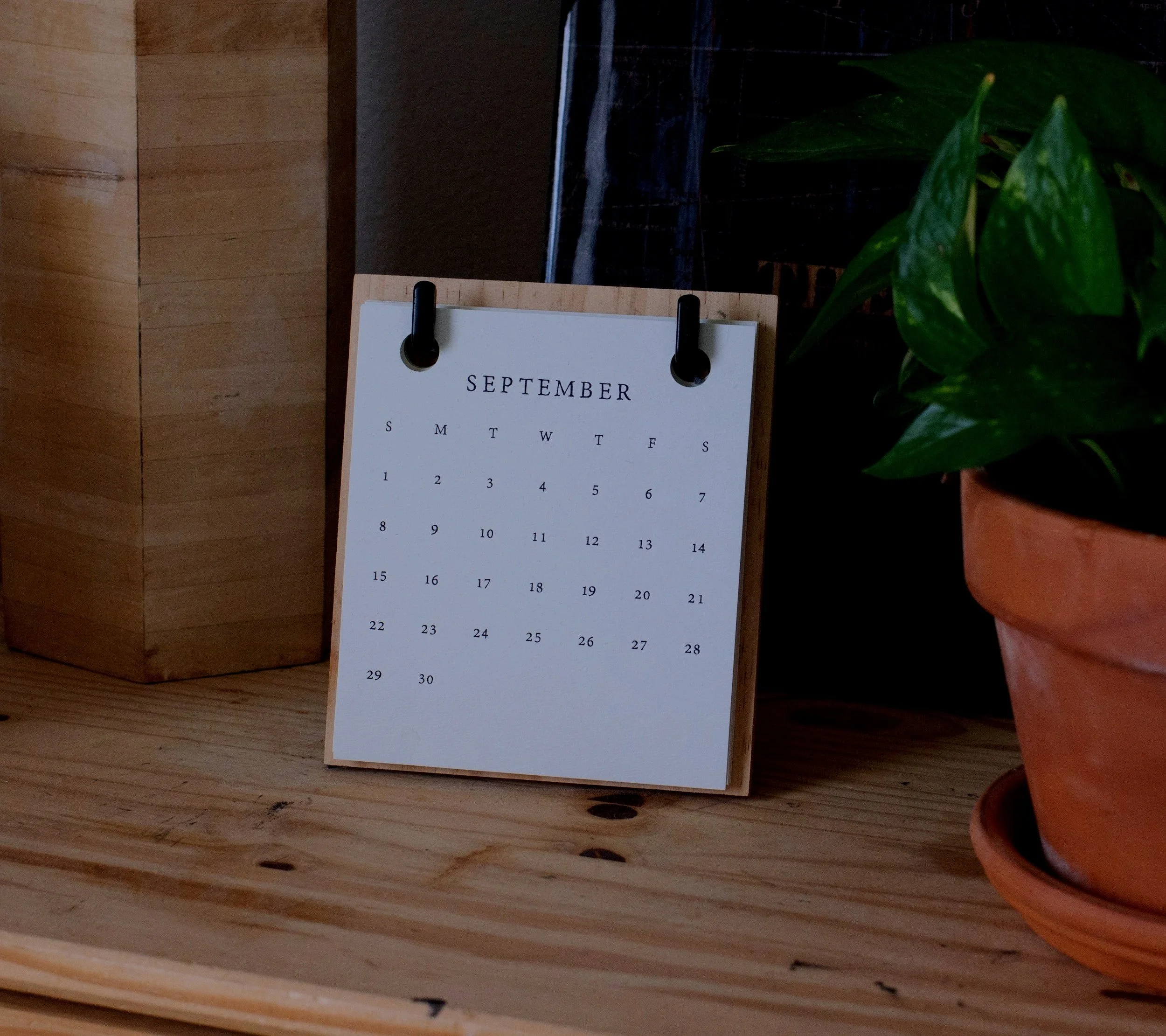 A small desk calendar displaying the month of September, placed on a wooden surface next to a potted green plant and a wooden box.