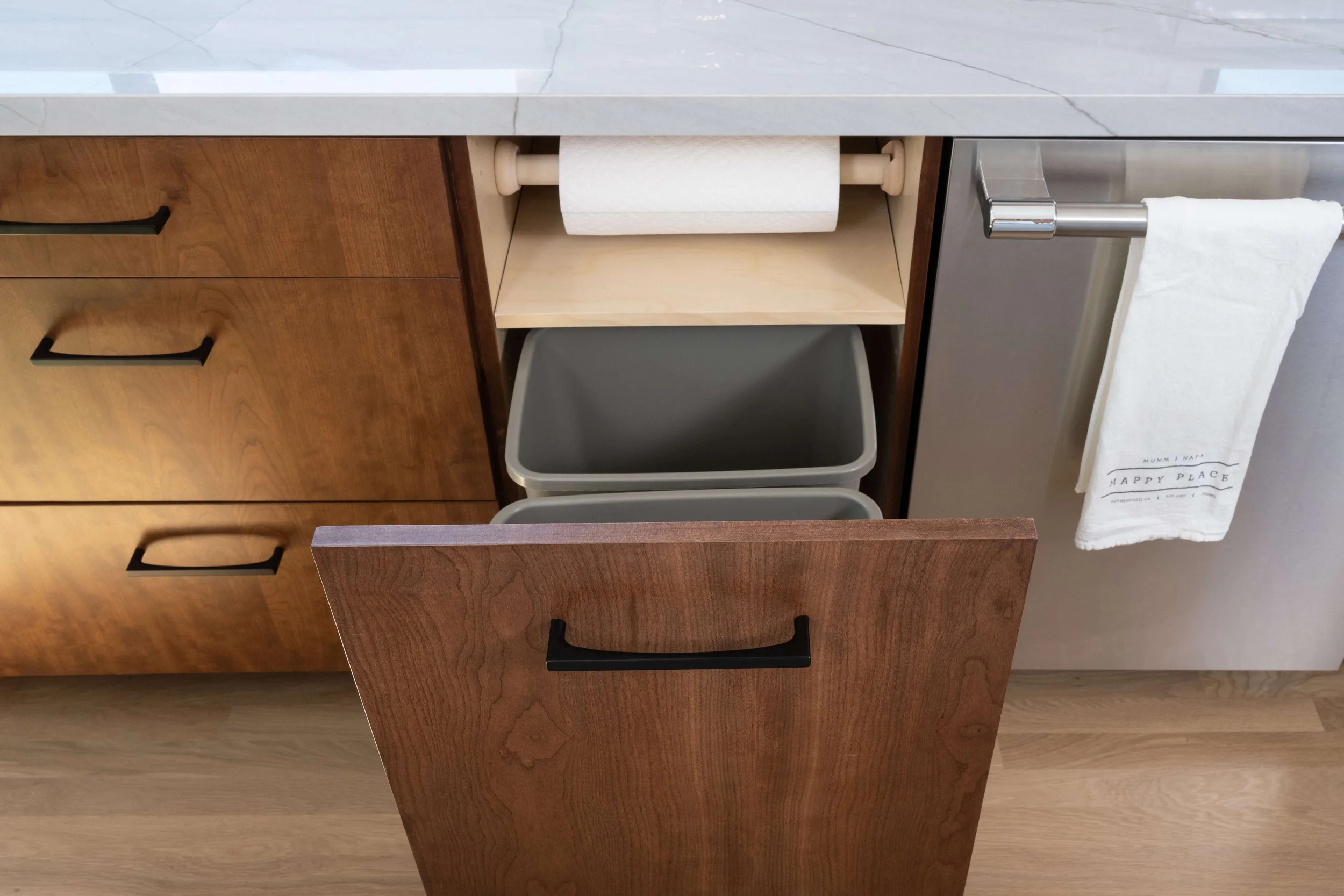 Open kitchen cabinet drawer with paper towel roll, trash bin, and a nearby hand towel with text 'HAPPY PLACE' hanging on the cabinet handle.