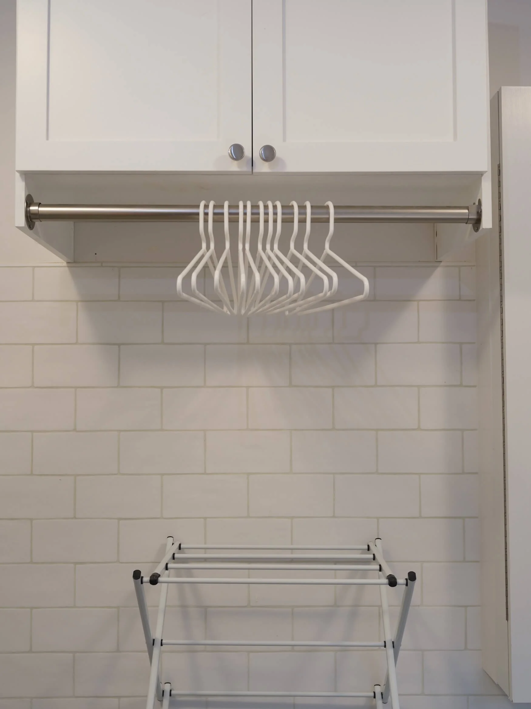 A white cabinet with two doors and round silver knobs, mounted on a white tiled wall. Below the cabinet, a stainless steel rod with white hangers is attached, and a collapsible white drying rack is positioned underneath.