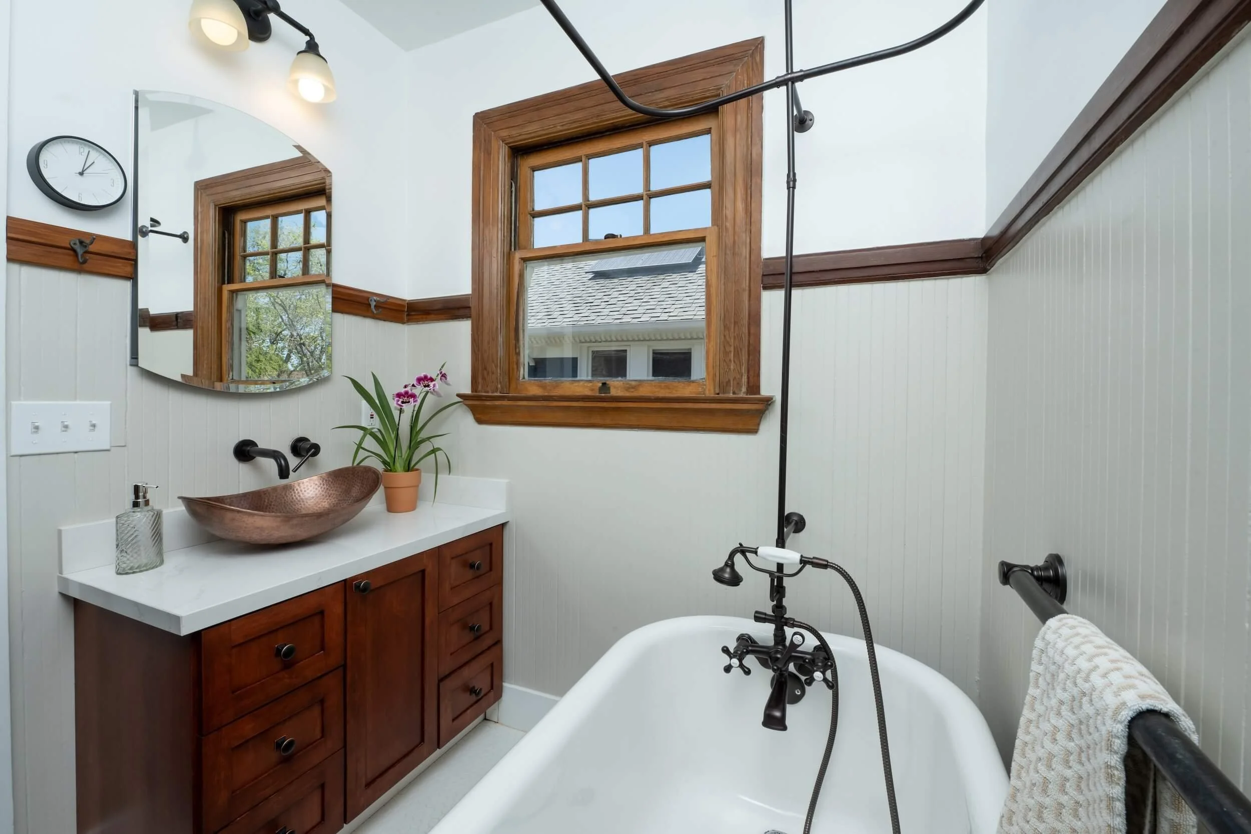 Bathroom with a clawfoot bathtub, black fixtures, and a wooden window with outside view. White beadboard walls, a vanity with a copper sink, mirror, potted plant, and wall clock.