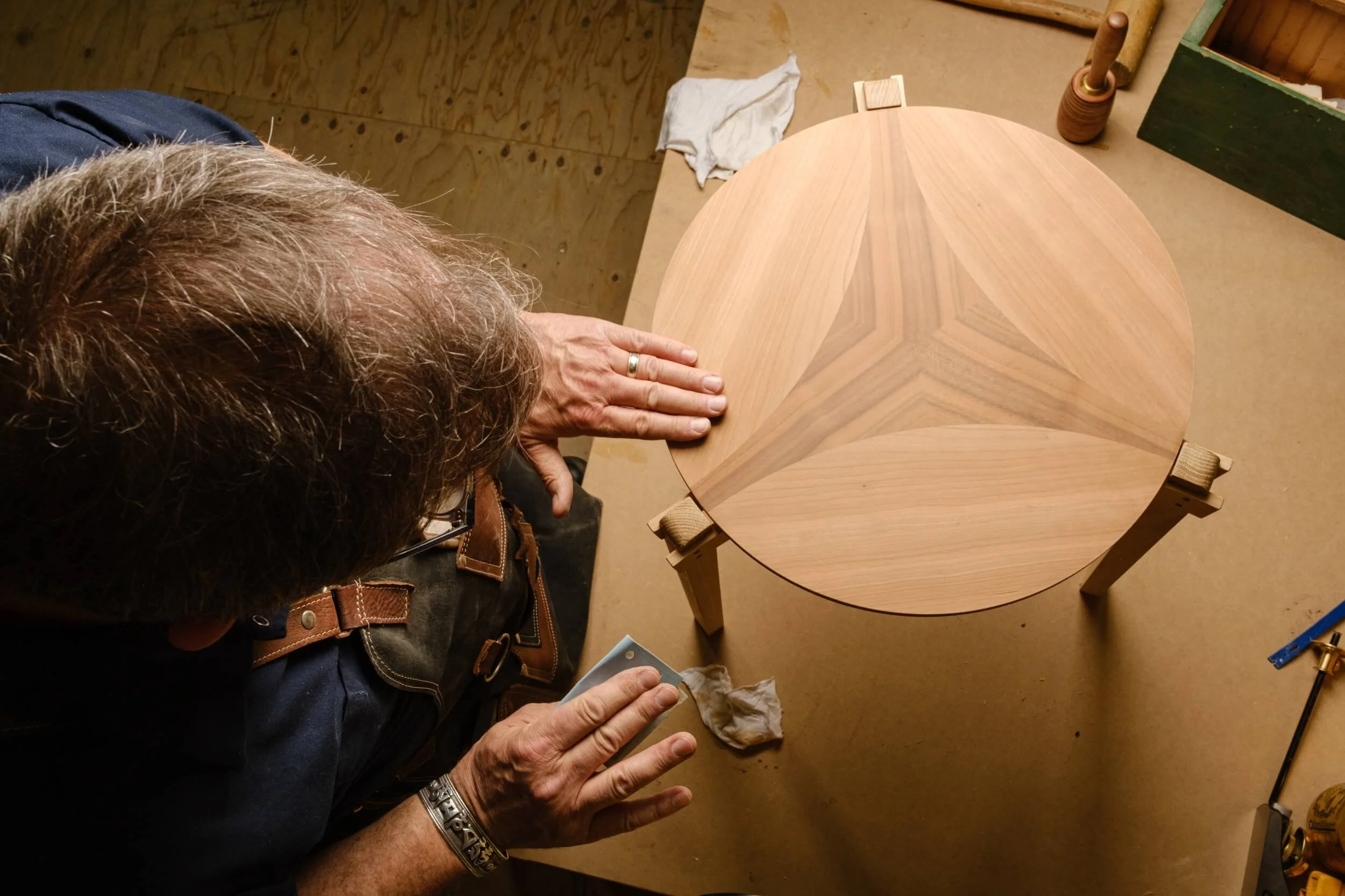 A person with long gray hair and a wedding band is working on a wooden table in a woodworking shop, with tools and a cloth nearby.
