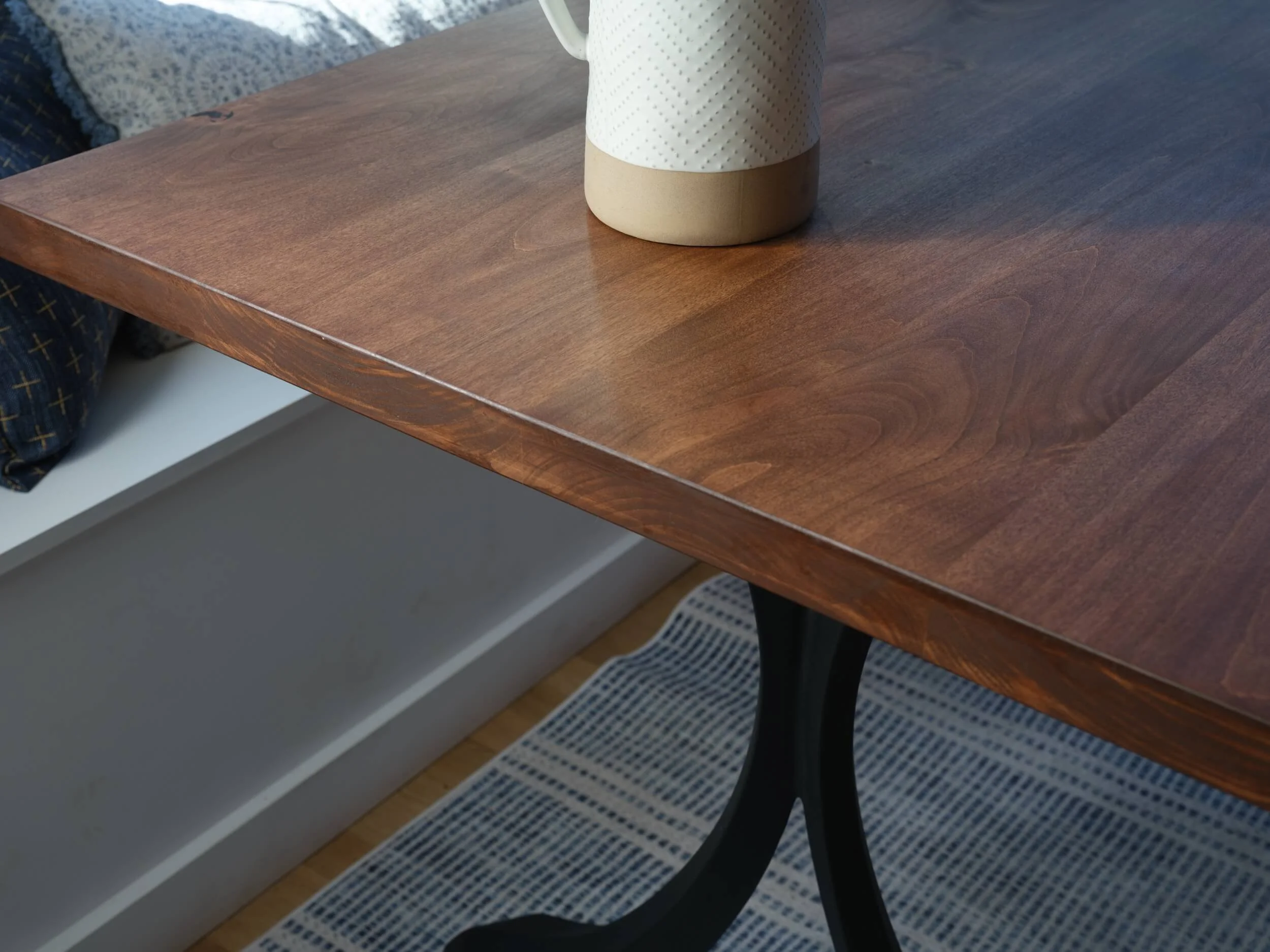 Close-up of a wooden table with a white ceramic mug featuring a textured pattern and a beige base. Part of a patterned cushion or blanket is visible in the background.