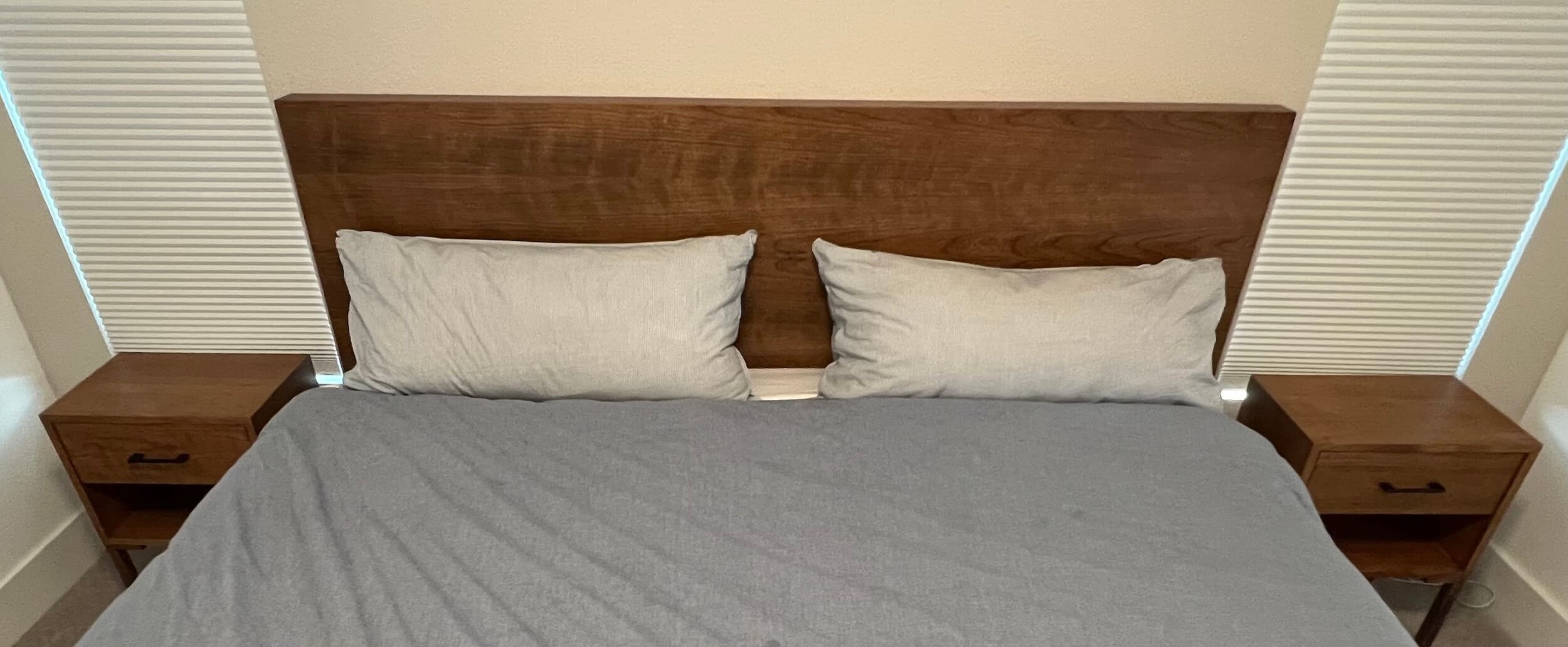 A neatly made bed with two beige pillows, a gray bedspread, and a wooden headboard with two matching bedside tables in a bedroom with beige walls and vertical blinds.
