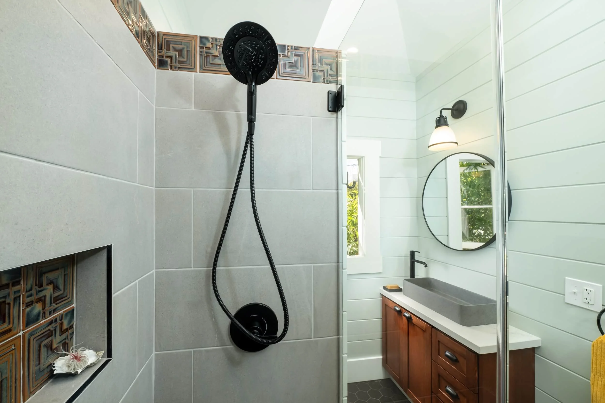 Bathroom with a black showerhead and hose on a tiled wall, a window, a mirror, a light fixture, a wooden vanity with a sink, and a yellow towel hanging on a rack.