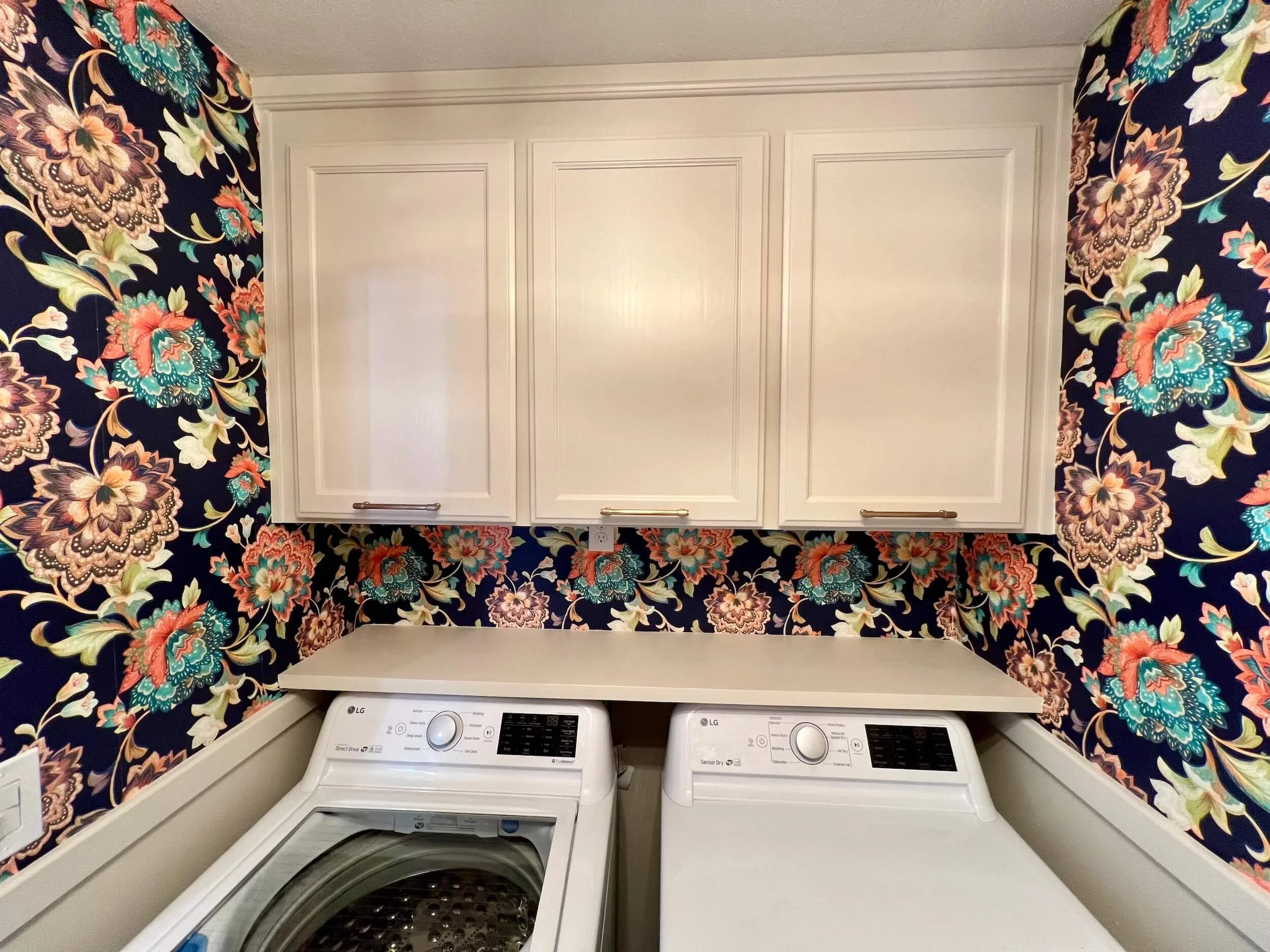 A laundry room with floral-patterned wallpaper and white cabinets above two white washing machines.