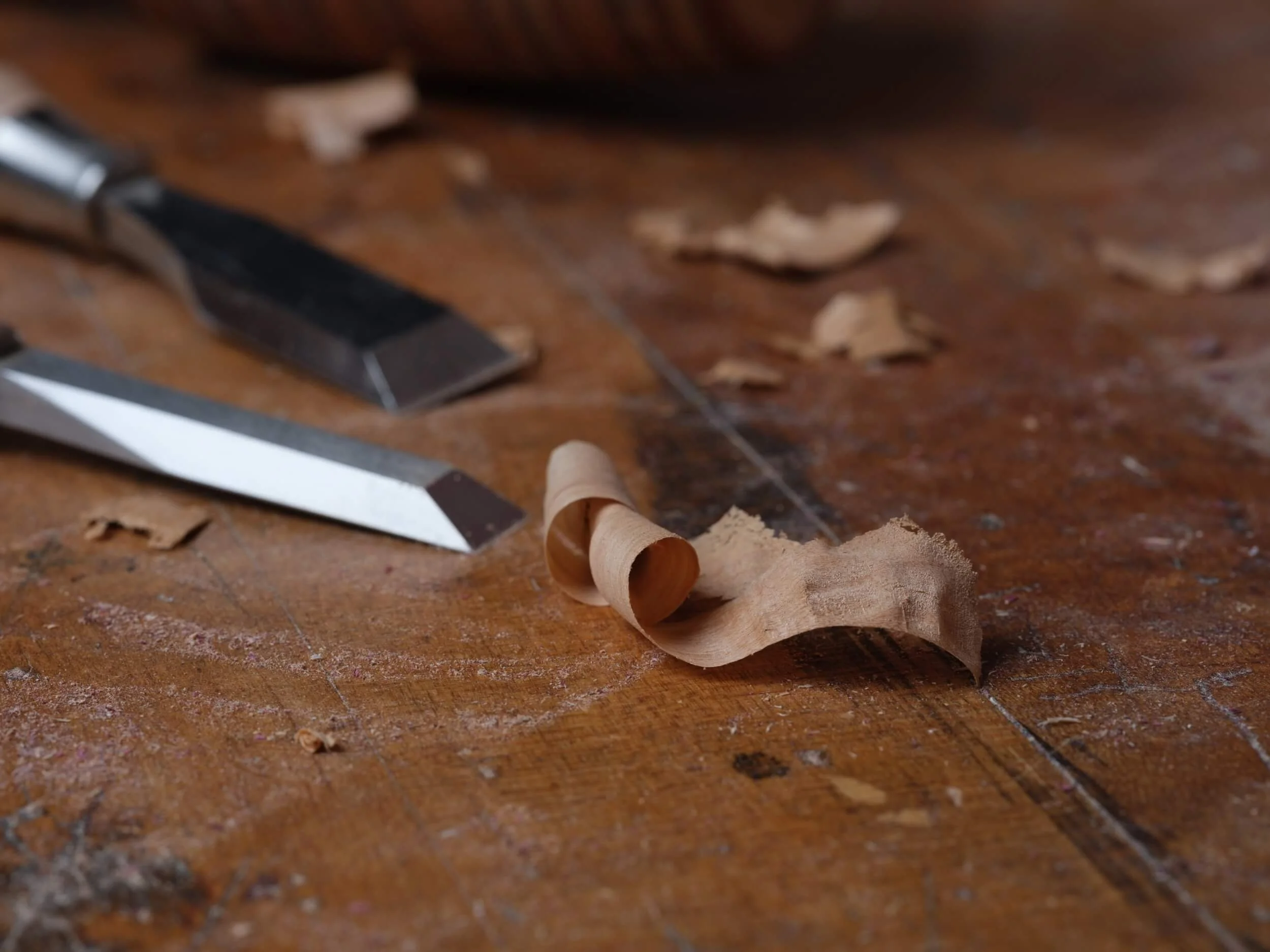 A wooden table with scattered paper shavings, a utility knife, and a small curled wood or paper strip.