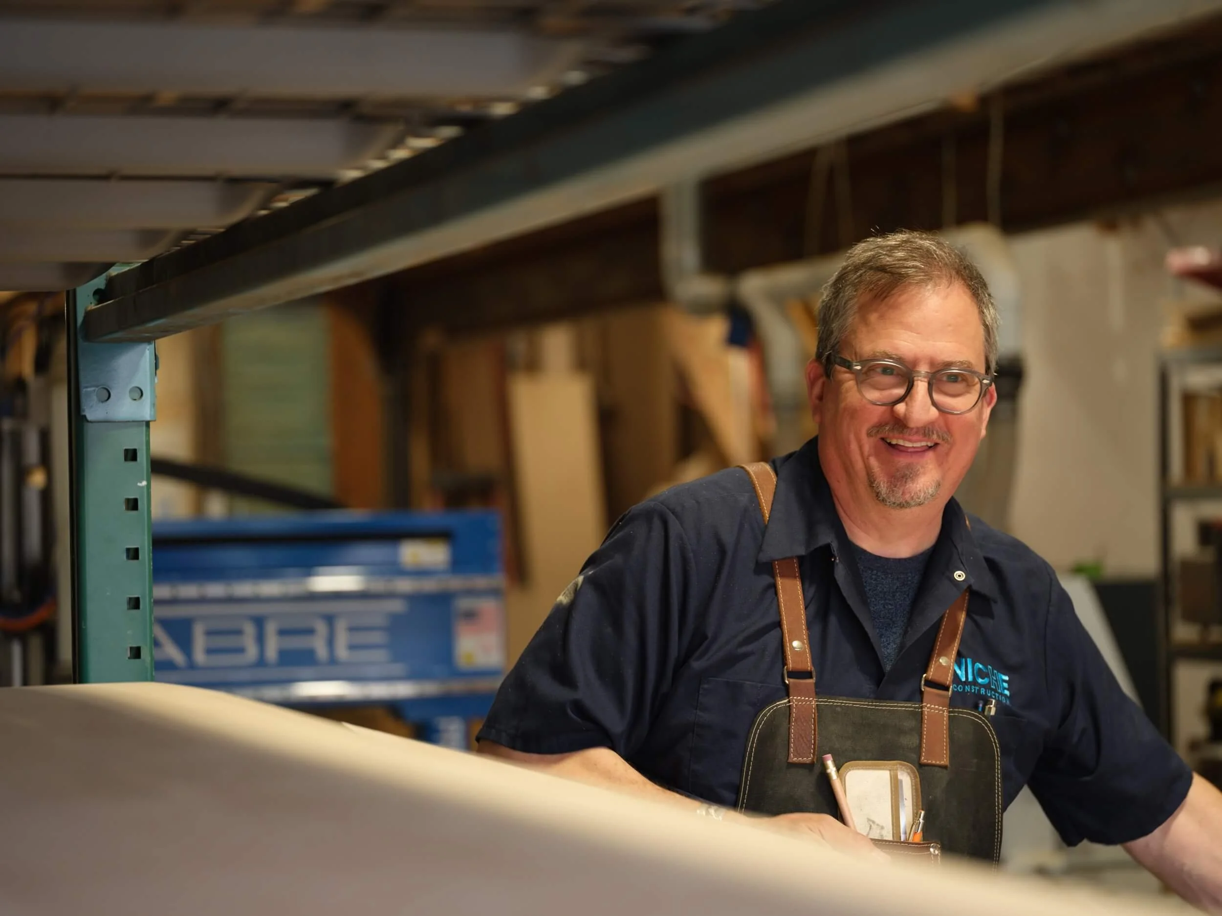 A man with glasses and a beard smiling at a construction site, wearing a dark shirt, leather apron, and a logo that says 'NICHE CONSTRUCTION'. There are shelves with tools and materials in the background.