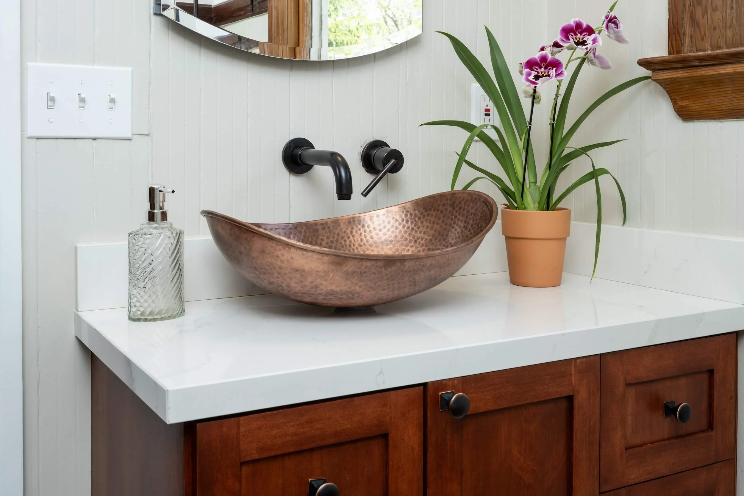 A bathroom sink area with a copper basin, a soap dispenser, a potted pink orchid plant, and a mirror above the sink. The background includes a white paneled wall and a wooden cabinet.