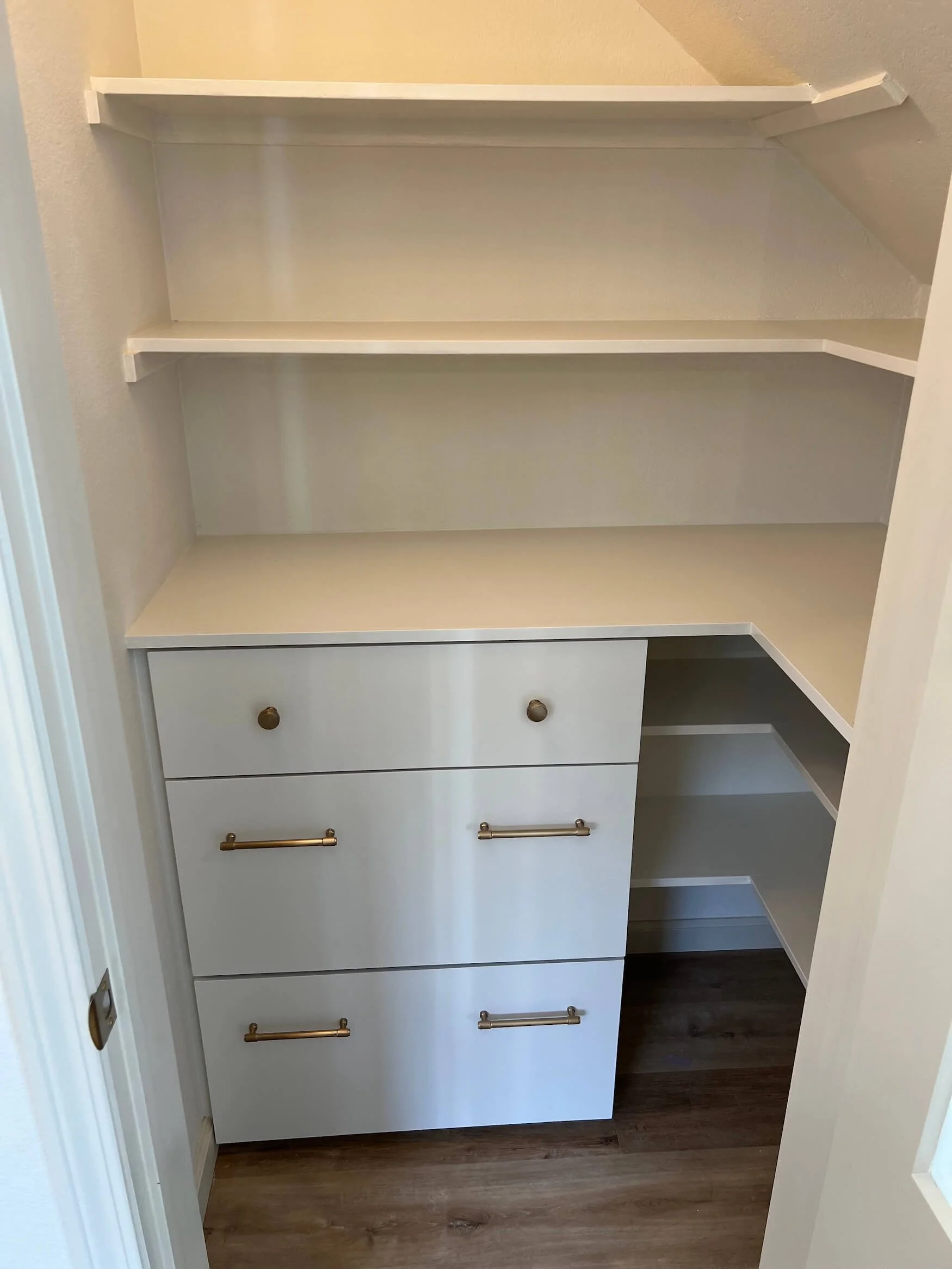 Empty white built-in shelves and drawers in a corner of a room with dark hardwood flooring.
