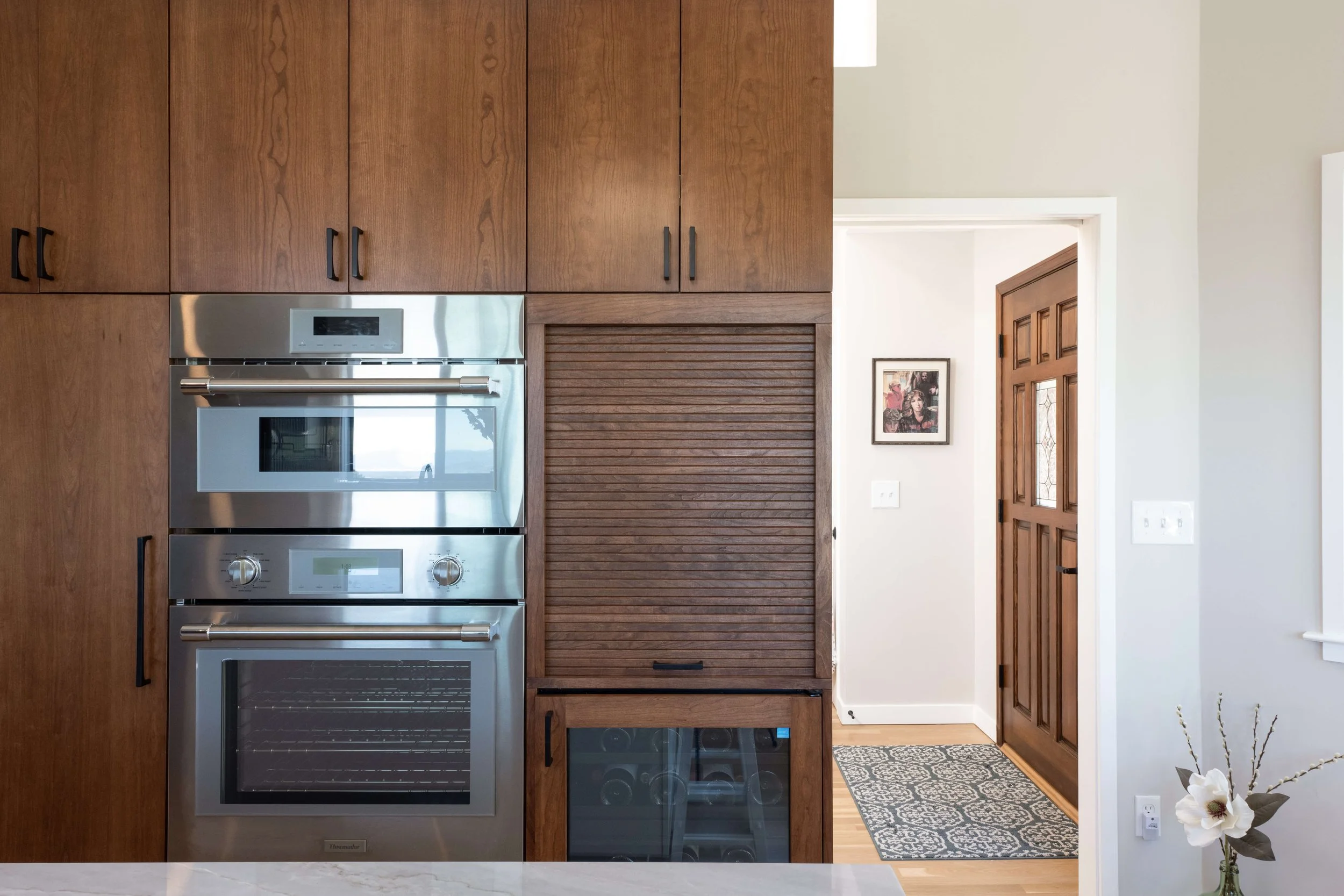 Interior view of a modern kitchen with wooden cabinets, stainless steel oven, and a doorway leading to an entryway with a wooden door and framed artwork.