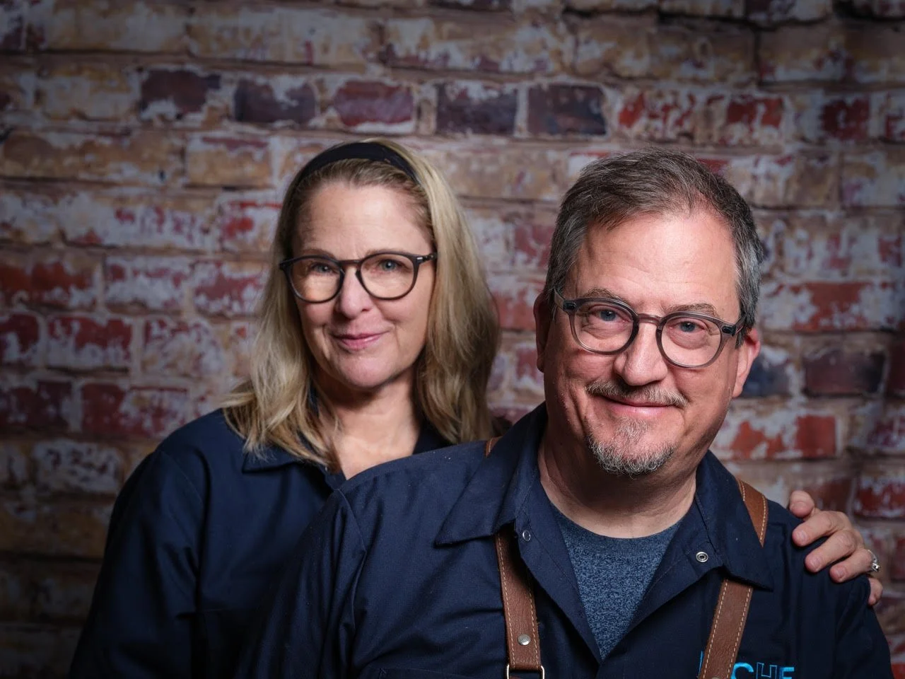 Portrait of a smiling middle-aged man and woman standing in front of a brick wall, both wearing glasses and dark shirts.