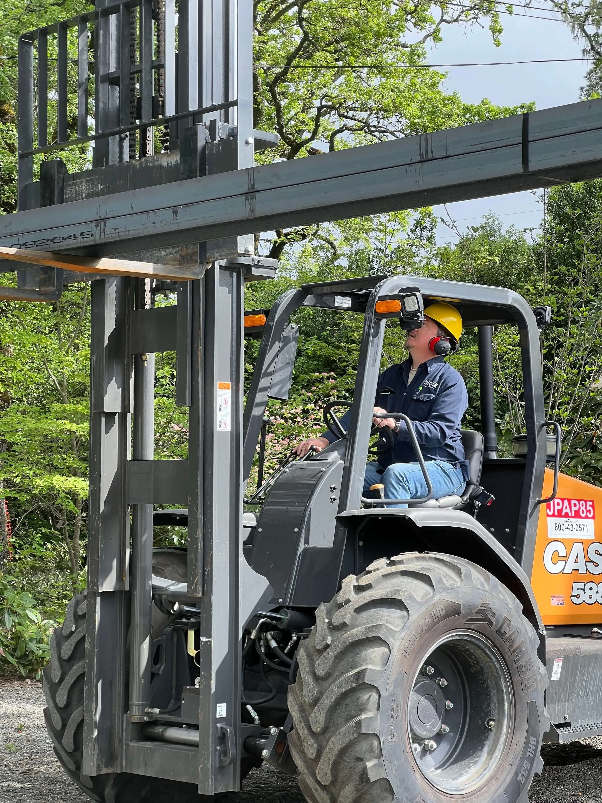 A worker operating a forklift in an outdoor area with green trees and shrubs behind, wearing a yellow helmet and safety ear protection.