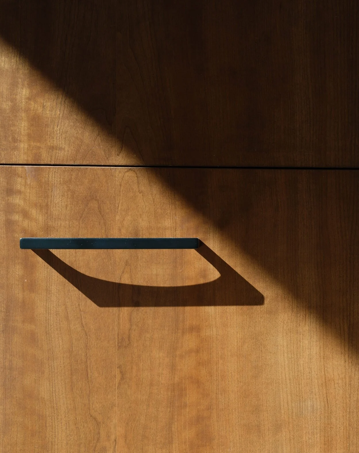 Close-up of a wooden cabinet or drawer with a black handle casting a shadow on the wood surface.