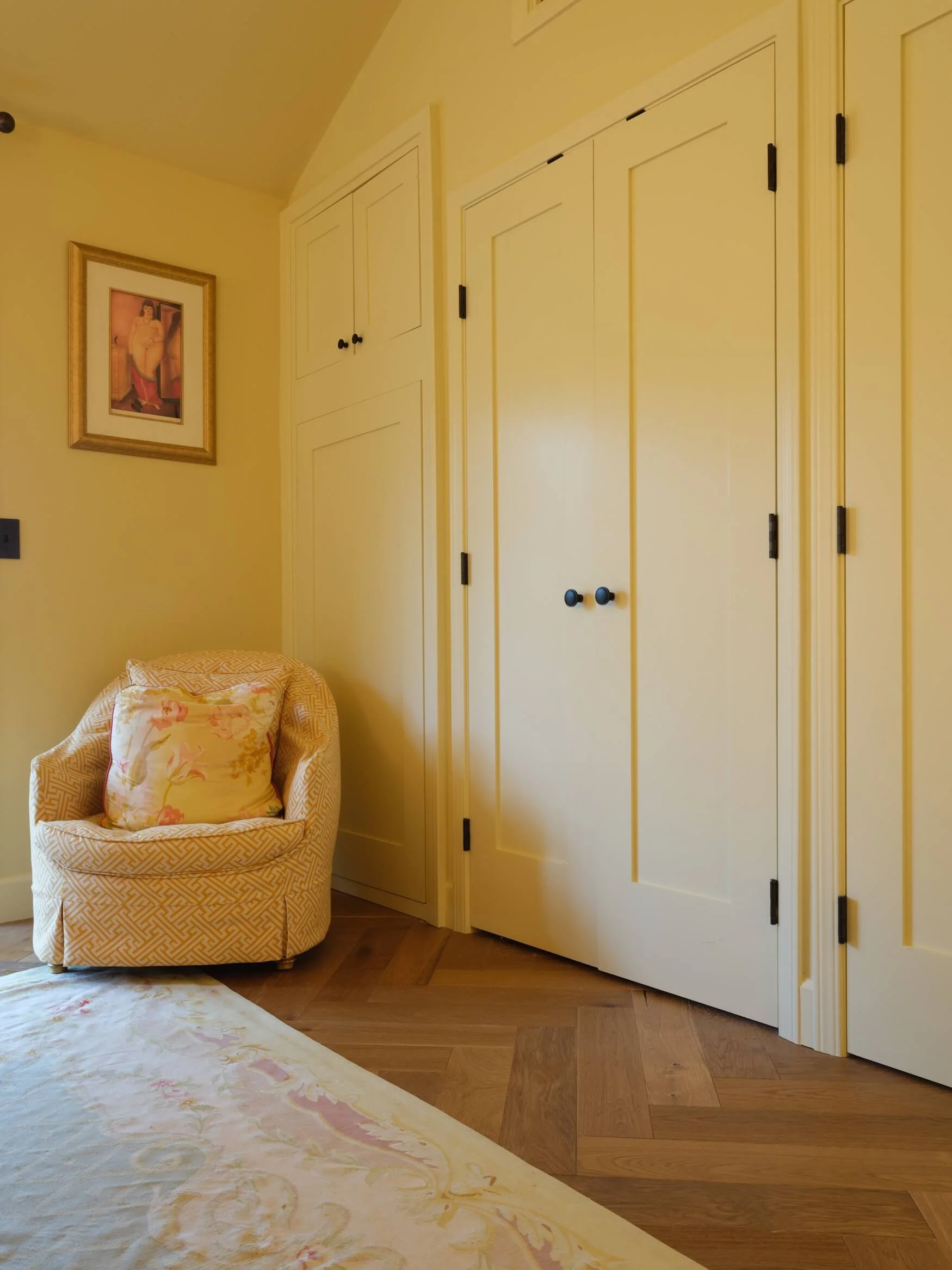 A corner of a room with yellow walls, a yellow armchair with a floral cushion, a wardrobe with multiple doors, and a framed artwork of a woman hanging on the wall.