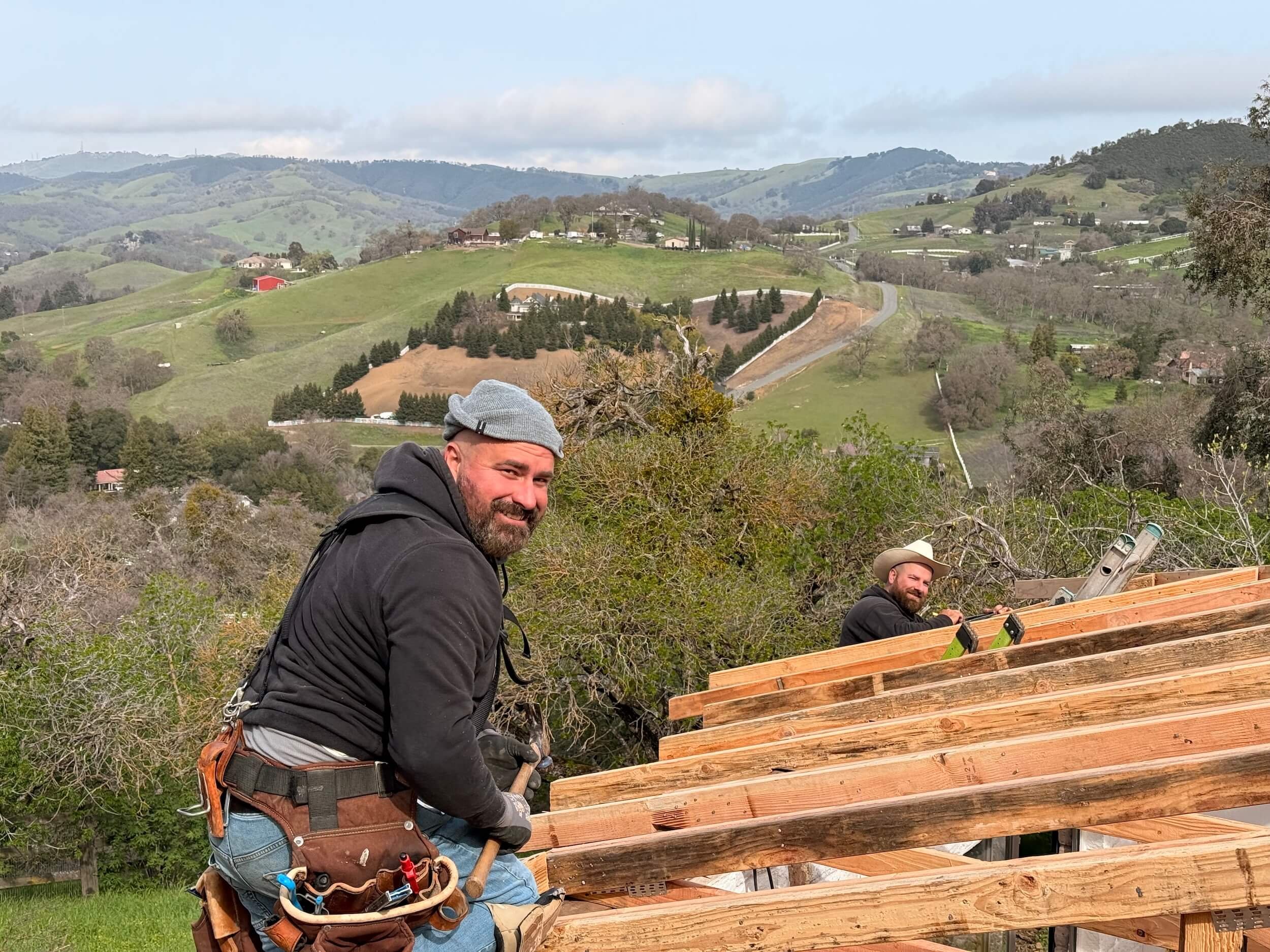 Two men building a wooden structure outdoors with a hilly landscape and houses in the background. One man is in black clothing and a gray beanie, and the other is wearing a beige hat.