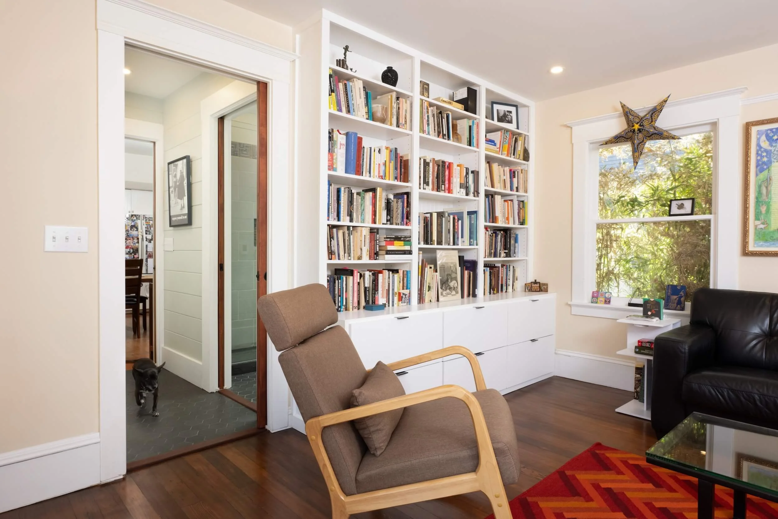A cozy living room with a large white bookshelf filled with books, a window with a star-shaped decoration, and a black leather sofa. A brown armchair and a red and orange patterned rug are also visible. A black and white photograph is on the bookshel