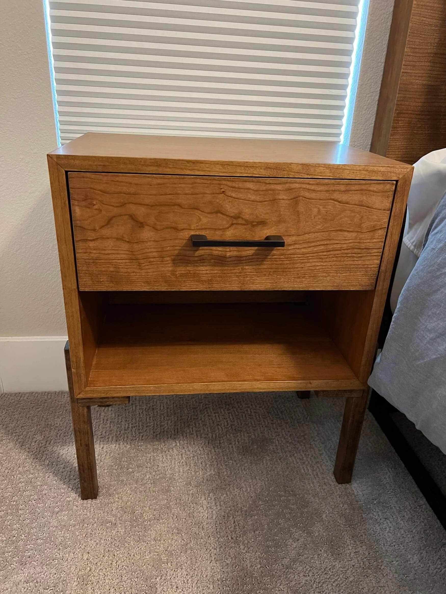 A wooden nightstand with a drawer and open shelf on tan carpet, positioned beside a bed with white bedding. A window with closed blinds is behind the nightstand.