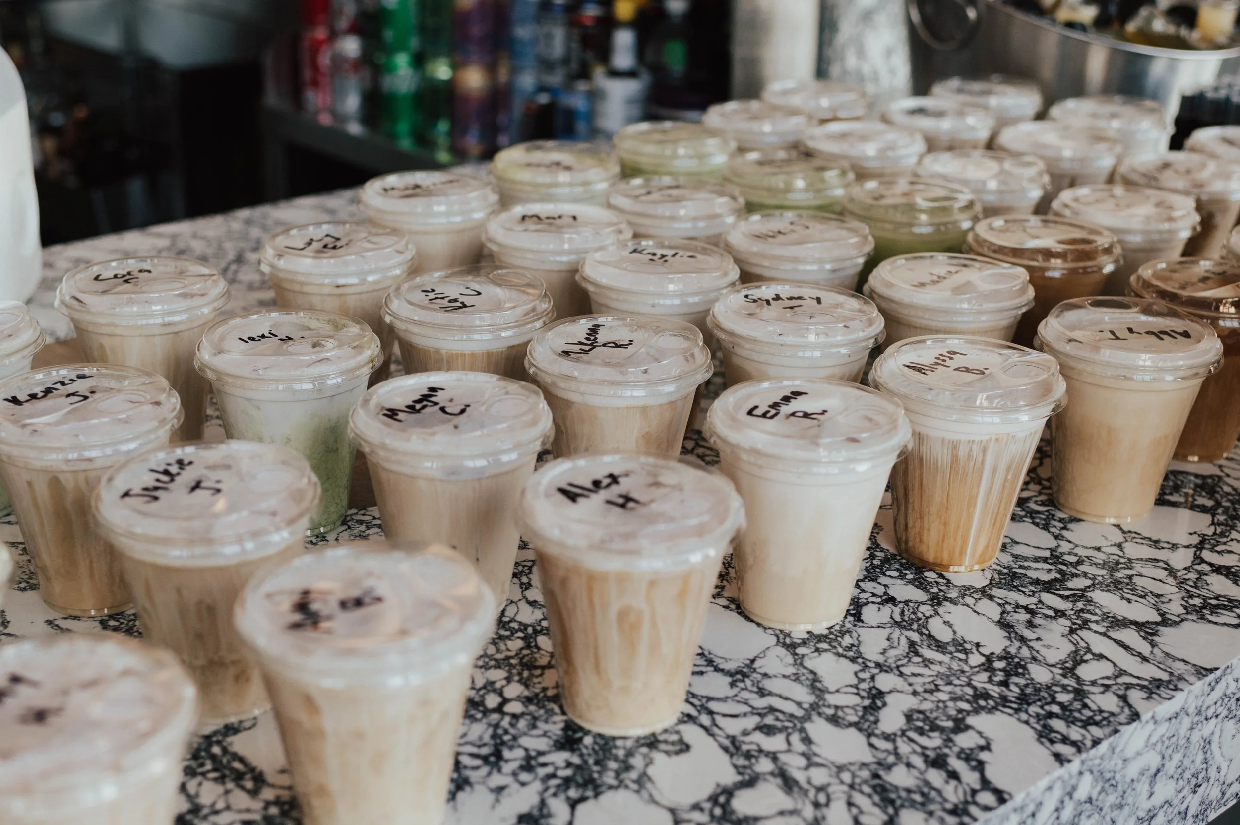A table filled with small cups of what appears to be ice cream or frozen dessert, each labeled with handwritten names on the lids. In the background, there are various bottles and containers.