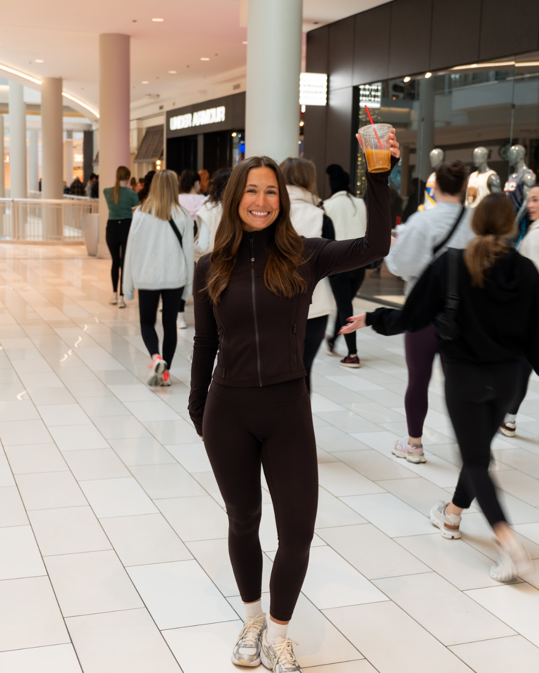 Smiling woman in workout clothes holding a cold beverage in a shopping mall.
