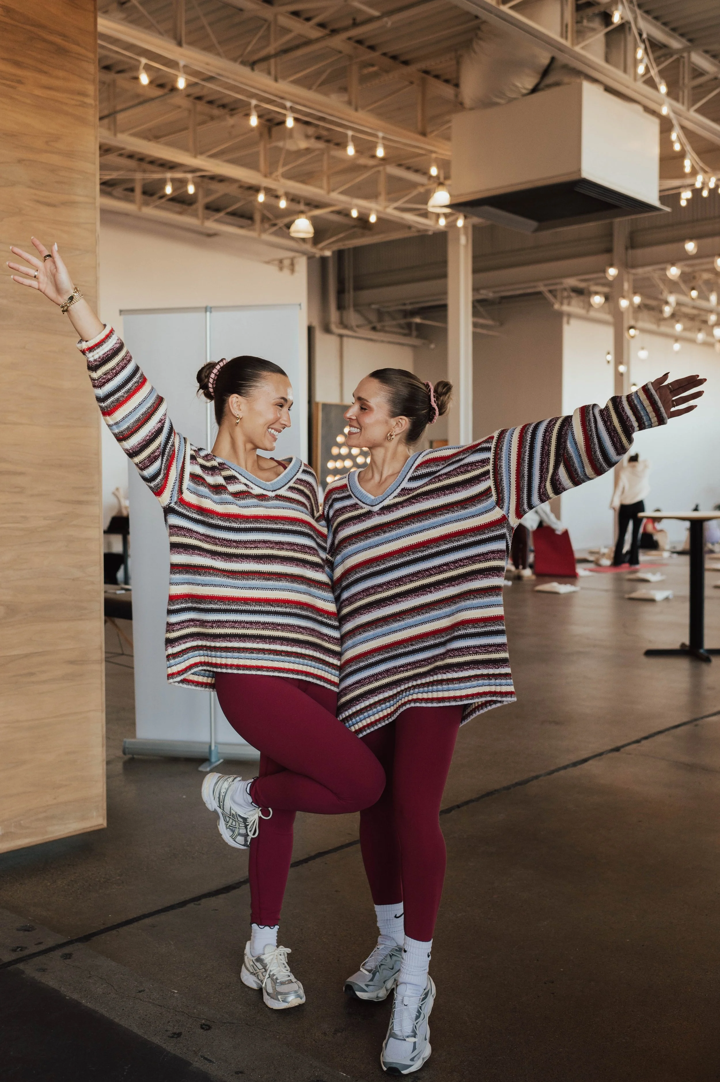 Two women wearing matching striped sweaters and maroon leggings, joyfully posing together indoors. One woman stands with her arm raised, the other kneels on one knee, smiling at each other.