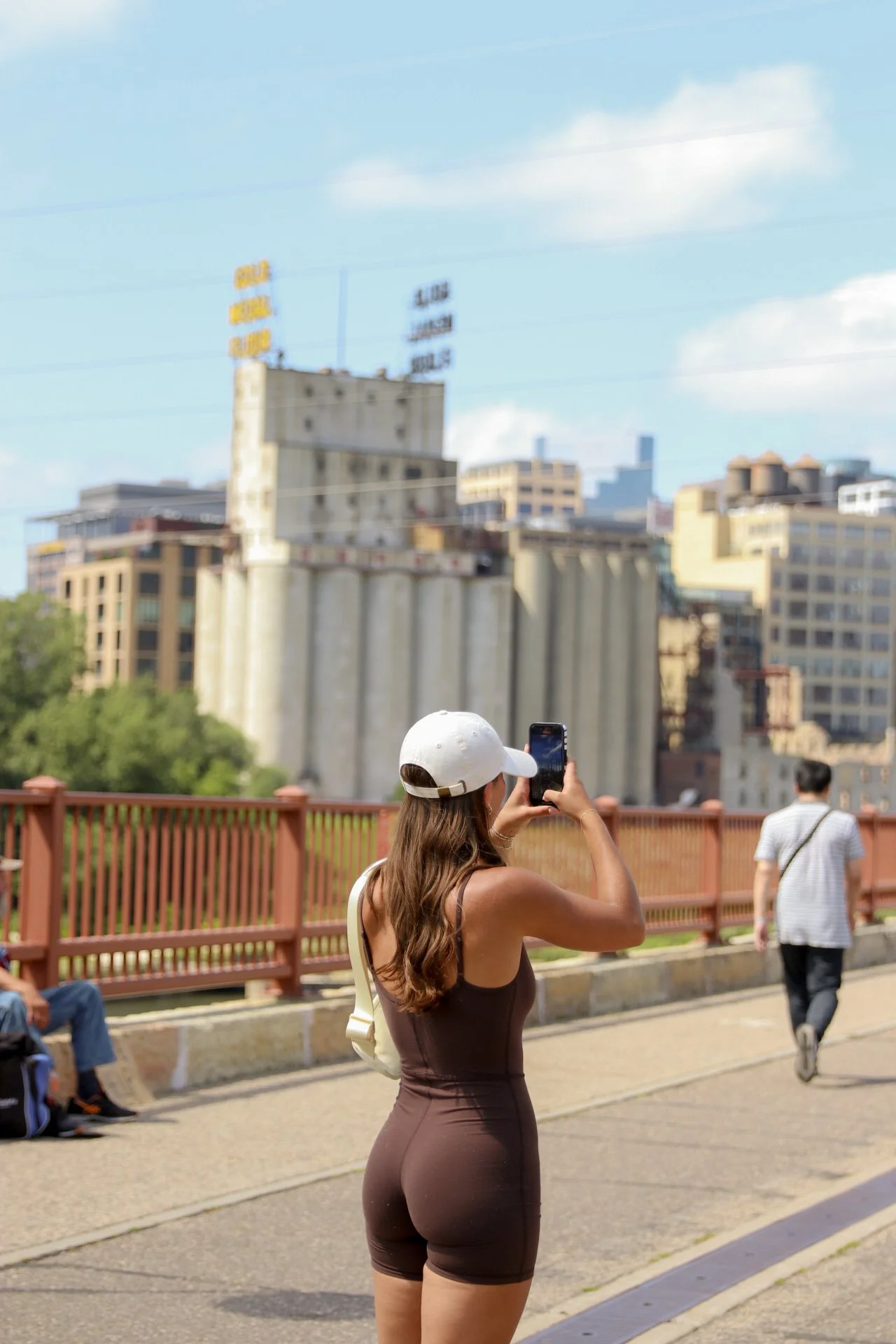 A woman taking a photo with her phone in an urban park, with buildings in the background and a person sitting on a bench to the left.