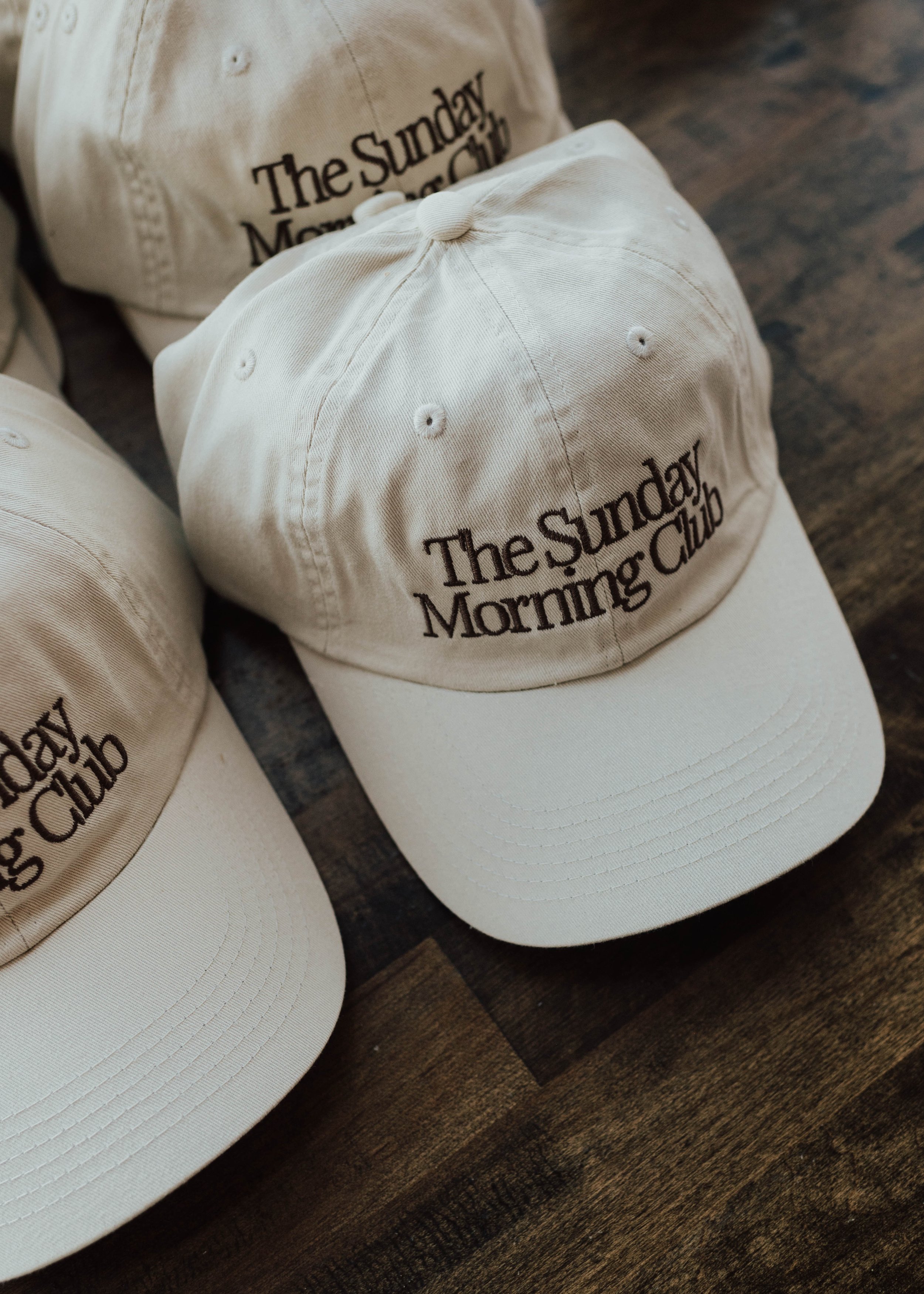 Several cream-colored baseball caps with the embroidered text "The Sunday Morning Club" on the front, placed on a wooden surface.