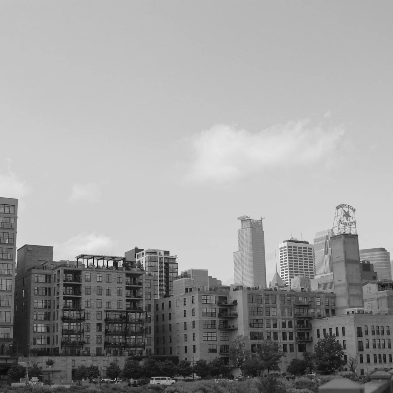 Black and white photo of a city skyline with a mix of tall modern skyscrapers and older mid-rise buildings, some with balconies, under a mostly clear sky with a few clouds.