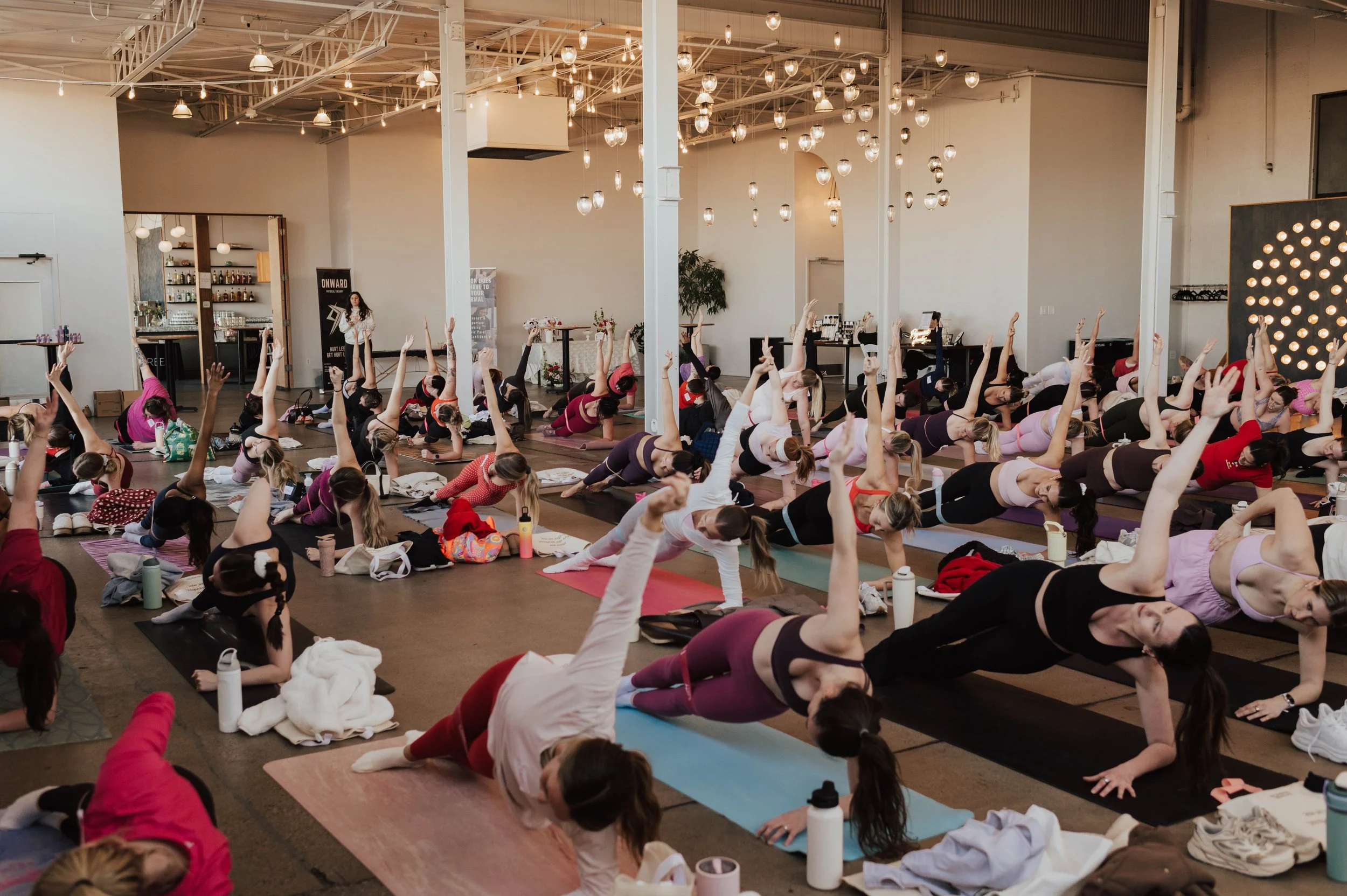 A large group of women participating in a yoga class or workshop in an indoor studio with high ceilings, hanging lights, and decorative lighting fixtures, all lying on yoga mats and extending their arms and legs in a stretching pose.