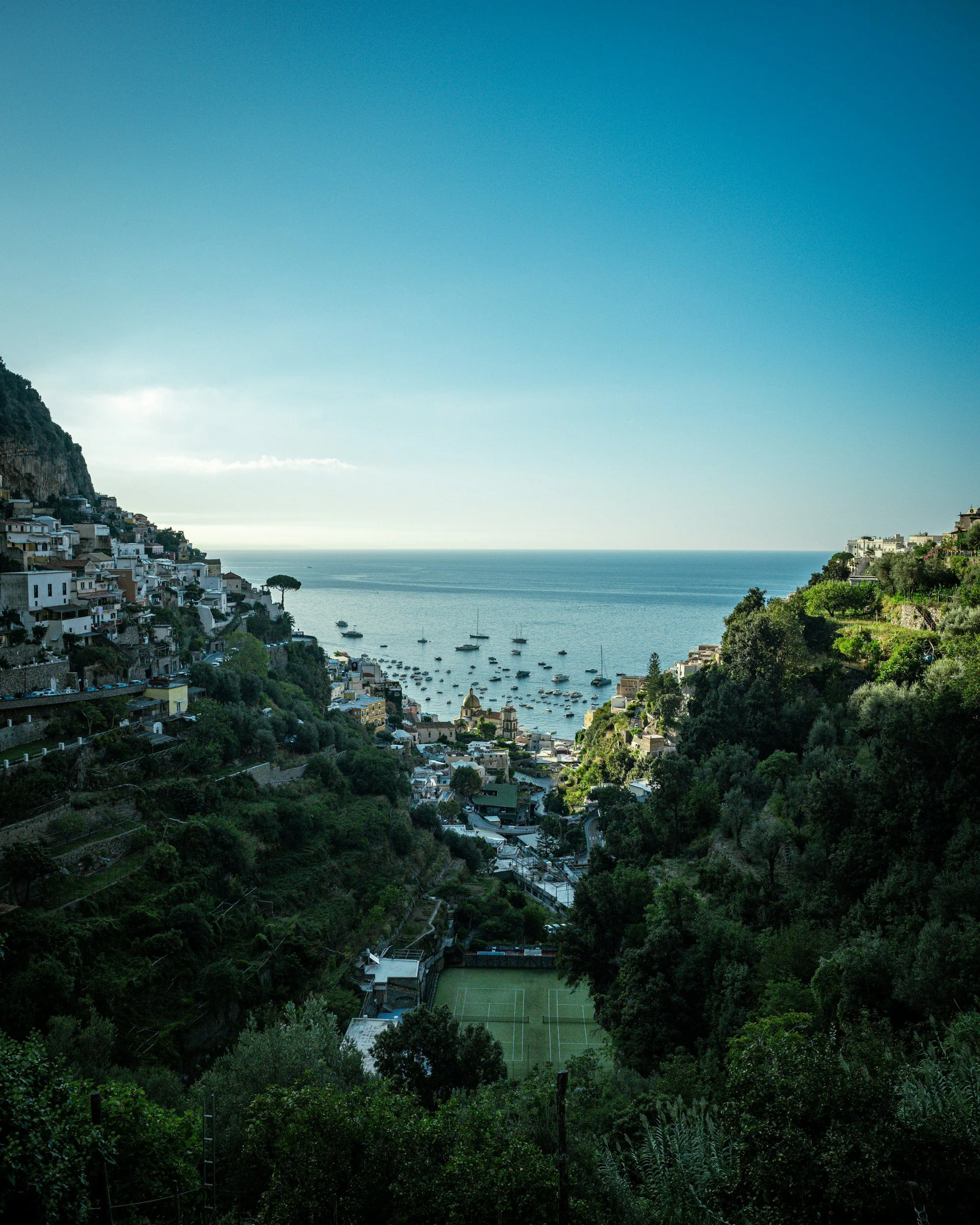 Scenic view of a coastal hillside town with houses, trees, a sports field, and boats in the bay, under a clear blue sky.