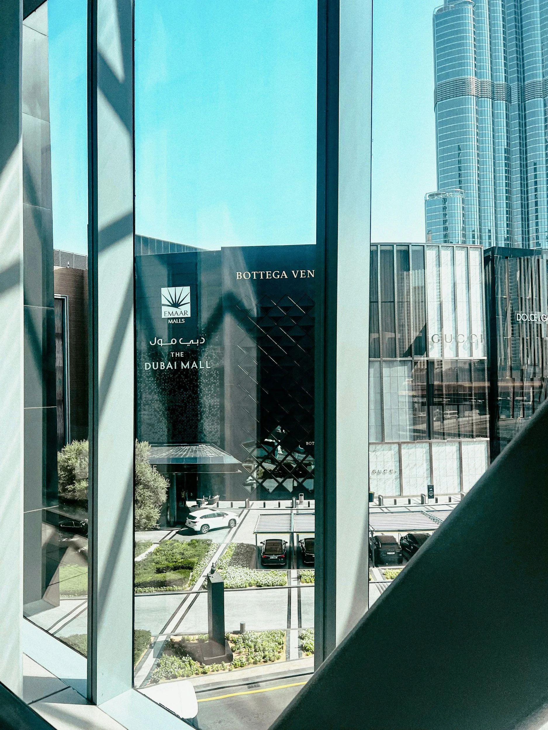 View of cityscape through glass windows showing luxury shopping mall signs for Bottega Veneta, Gucci, Dolce & Gabbana, and The Dubai Mall.
