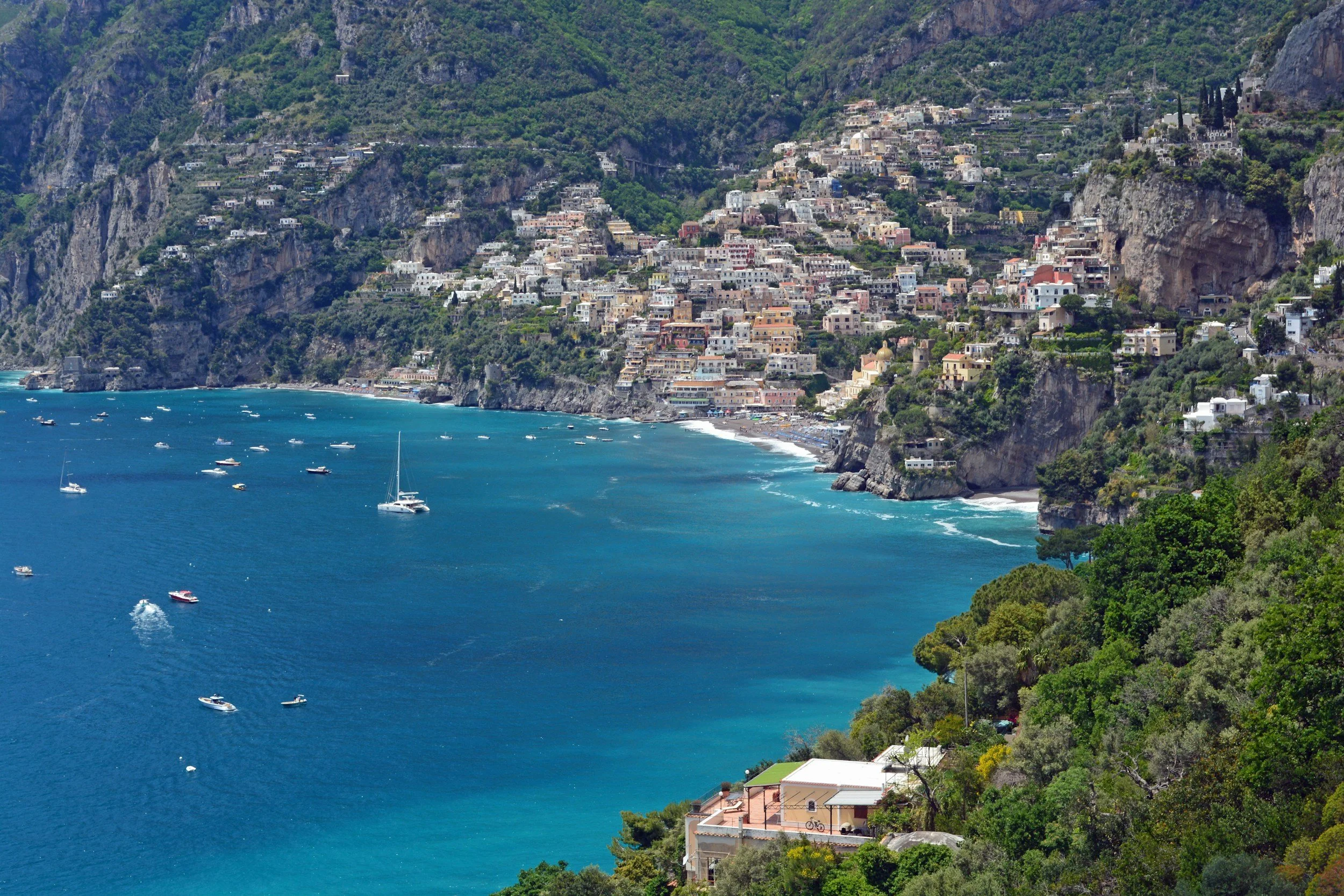 The image shows a coastal hillside town with colorful buildings stacked on the steep slopes, overlooking the blue sea with boats.