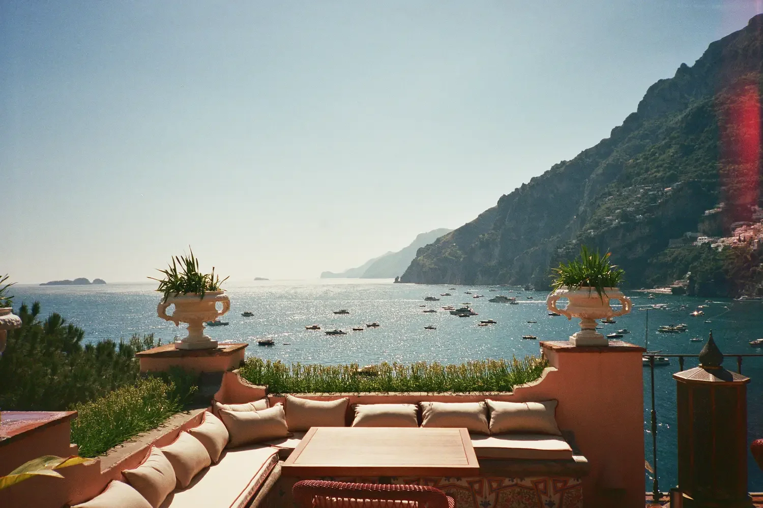 View of a terrace with cushioned seating overlooking a bayside with boats, mountains, and clear sky.