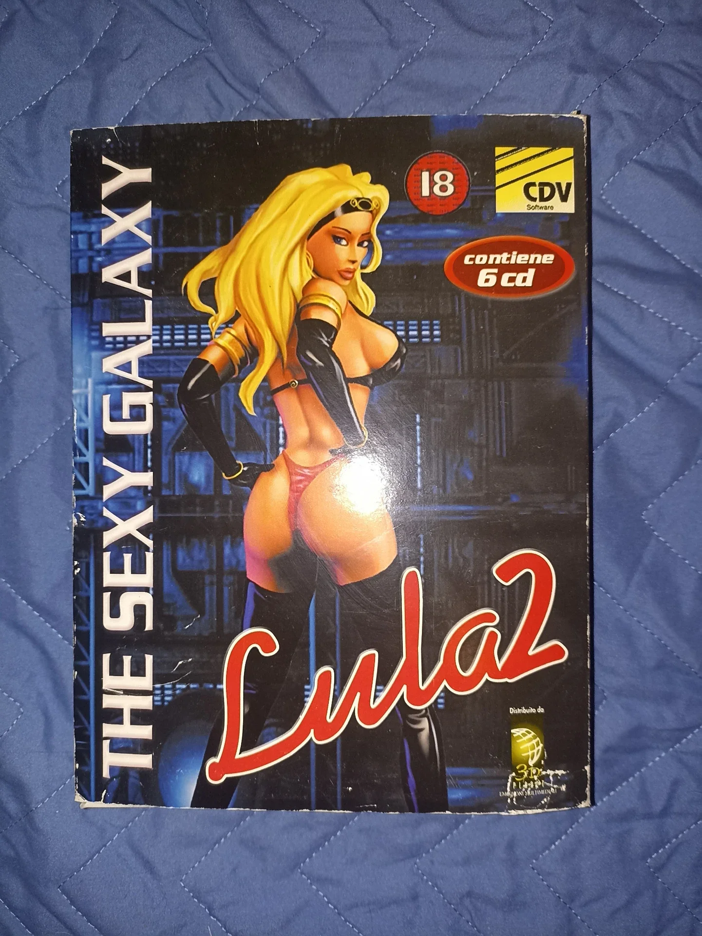 Copertina di un DVD con un'illustrazione di una donna con capelli biondi lunghi, vestita con abiti succinti e stivali alti, in un ambiente futuristico. Include vari loghi e informazioni, come l'età minima raccomandata di 18 anni e che contiene 6 CD.