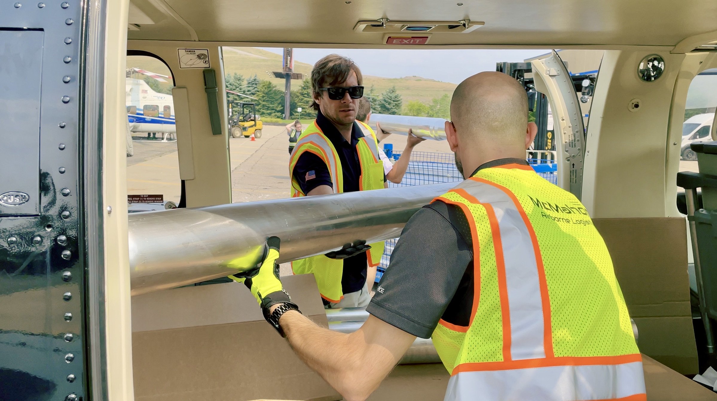Two men in high-visibility vests moving a large cylindrical object into an aircraft or helicopter. One man is wearing sunglasses and a black shirt, while the other has a shaved head. In the background, there are vehicles, a forklift, and a hilly landscape.