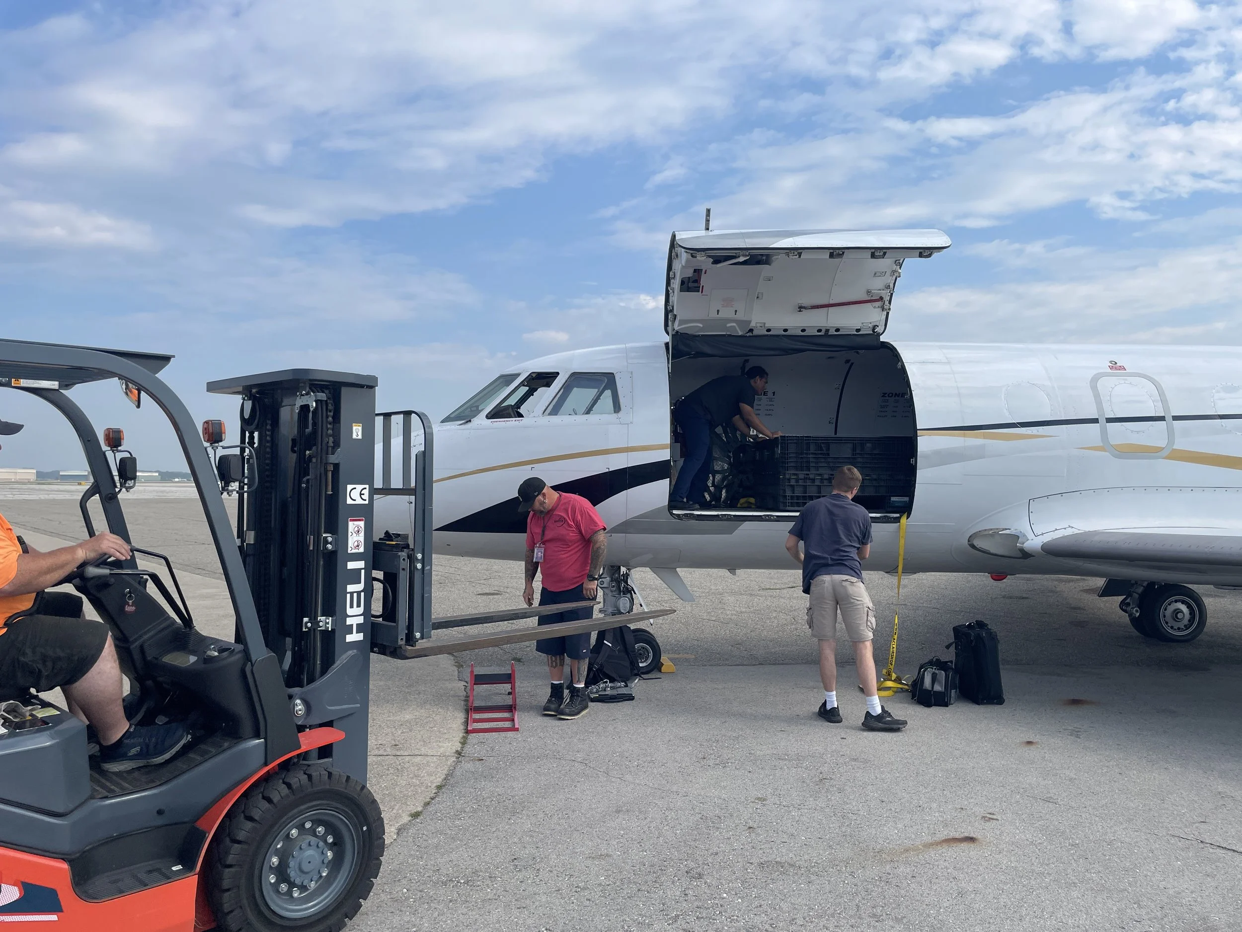 Ground crew loading luggage into the cargo hold of a white commercial airplane on an airport tarmac. Several ground staff are working, some standing near the aircraft with luggage, and a forklift is visible in the foreground.