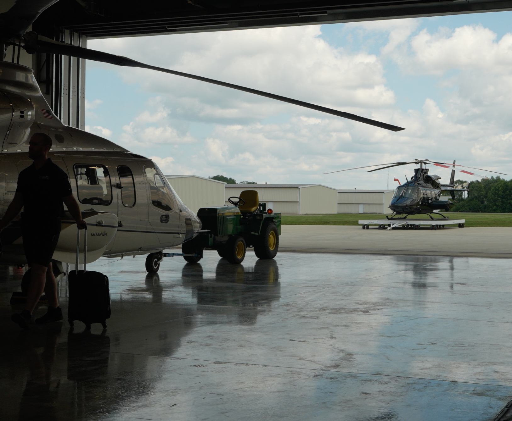 Inside an airplane hangar, a person is walking with luggage. A helicopter is parked outside on the tarmac, while another helicopter is still inside the hangar.