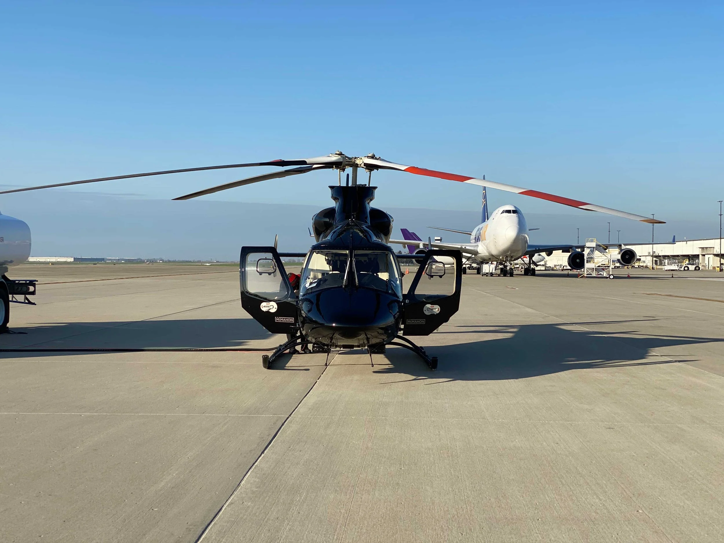 A black helicopter parked on an airport tarmac with commercial airplanes in the background under a clear sky.