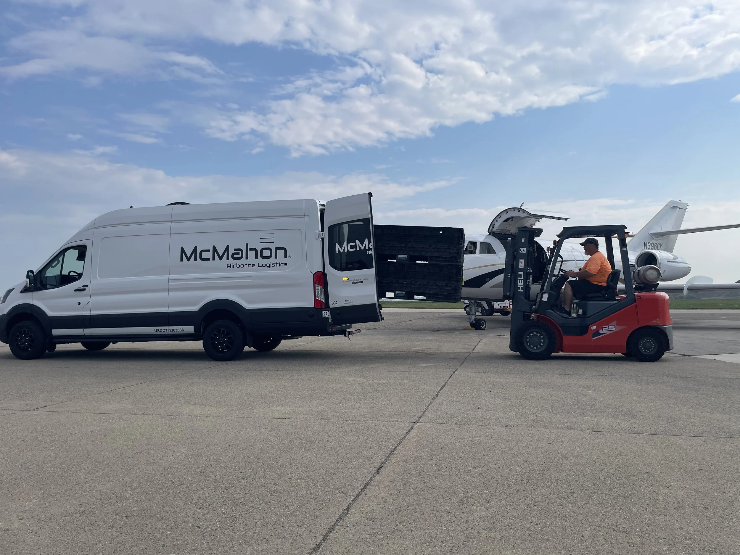 A man operating a forklift on an airport tarmac, moving a large black container near a small passenger jet airplane. There is also a white delivery van with the brand 'McMahon' parked nearby, and the sky is partly cloudy.