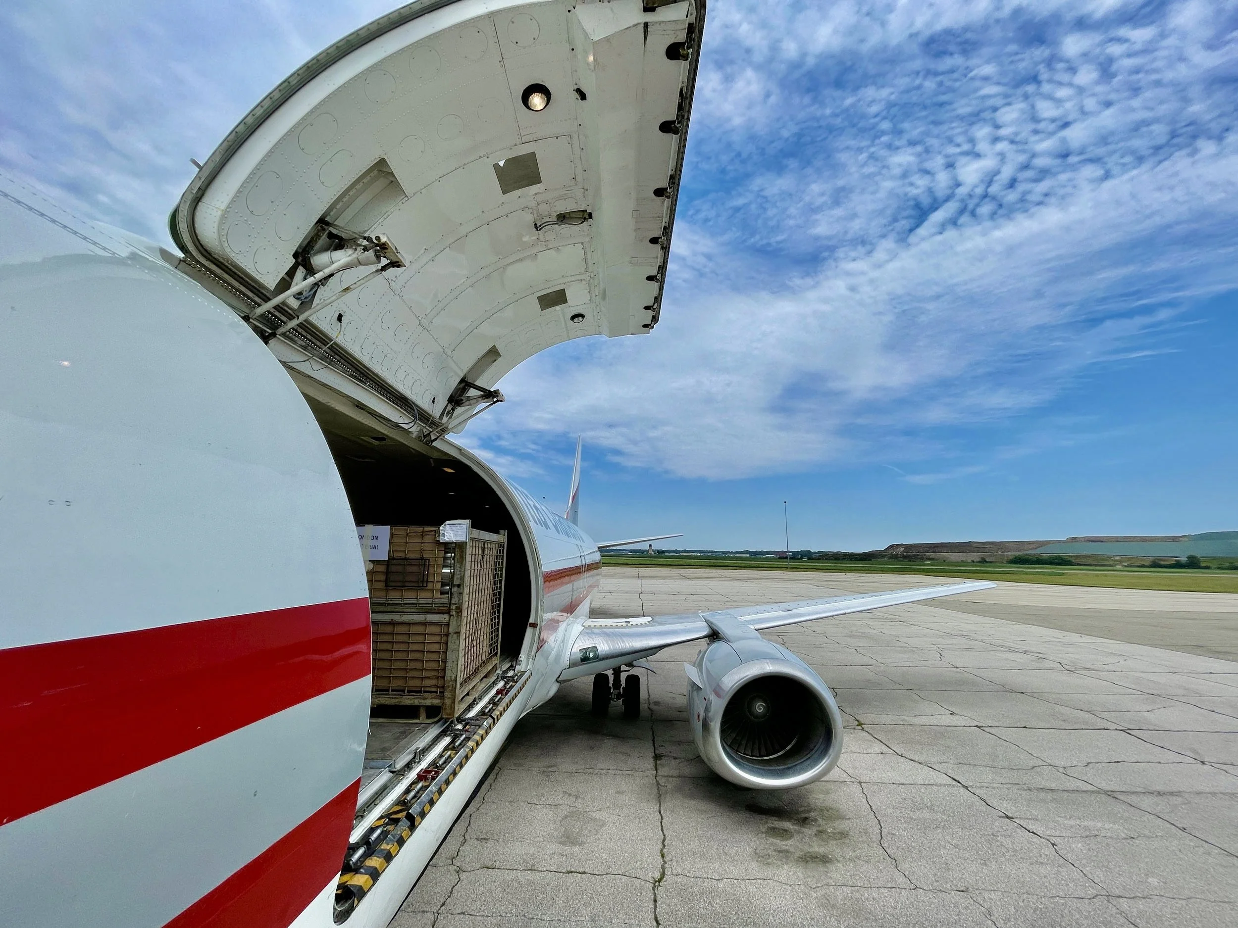 View of an airplane at the airport with luggage being loaded into the cargo hold, under a partly cloudy sky.