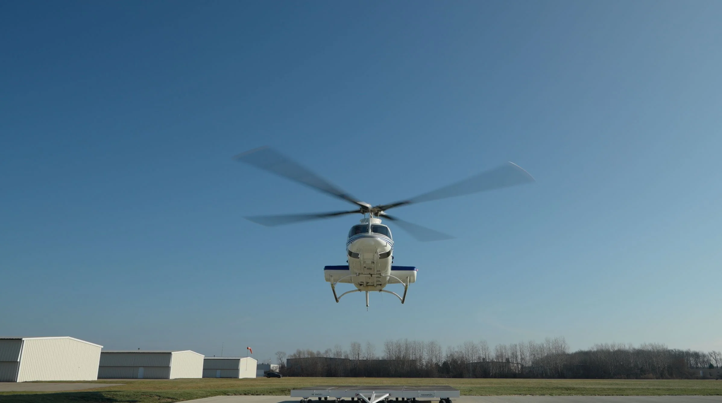 A white helicopter flying in clear blue sky over an open area with trees and buildings in the background.