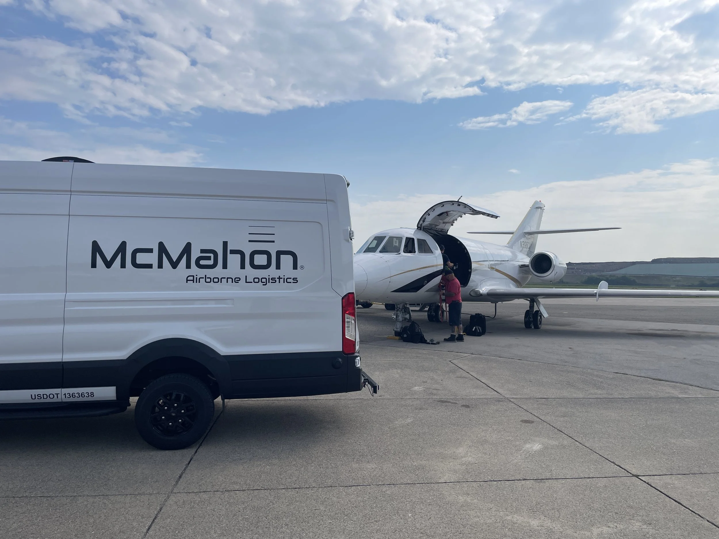 A private jet with its door open and a person standing beside it on the tarmac, with a McMahon Airborne Logistics truck nearby and a partly cloudy sky overhead.